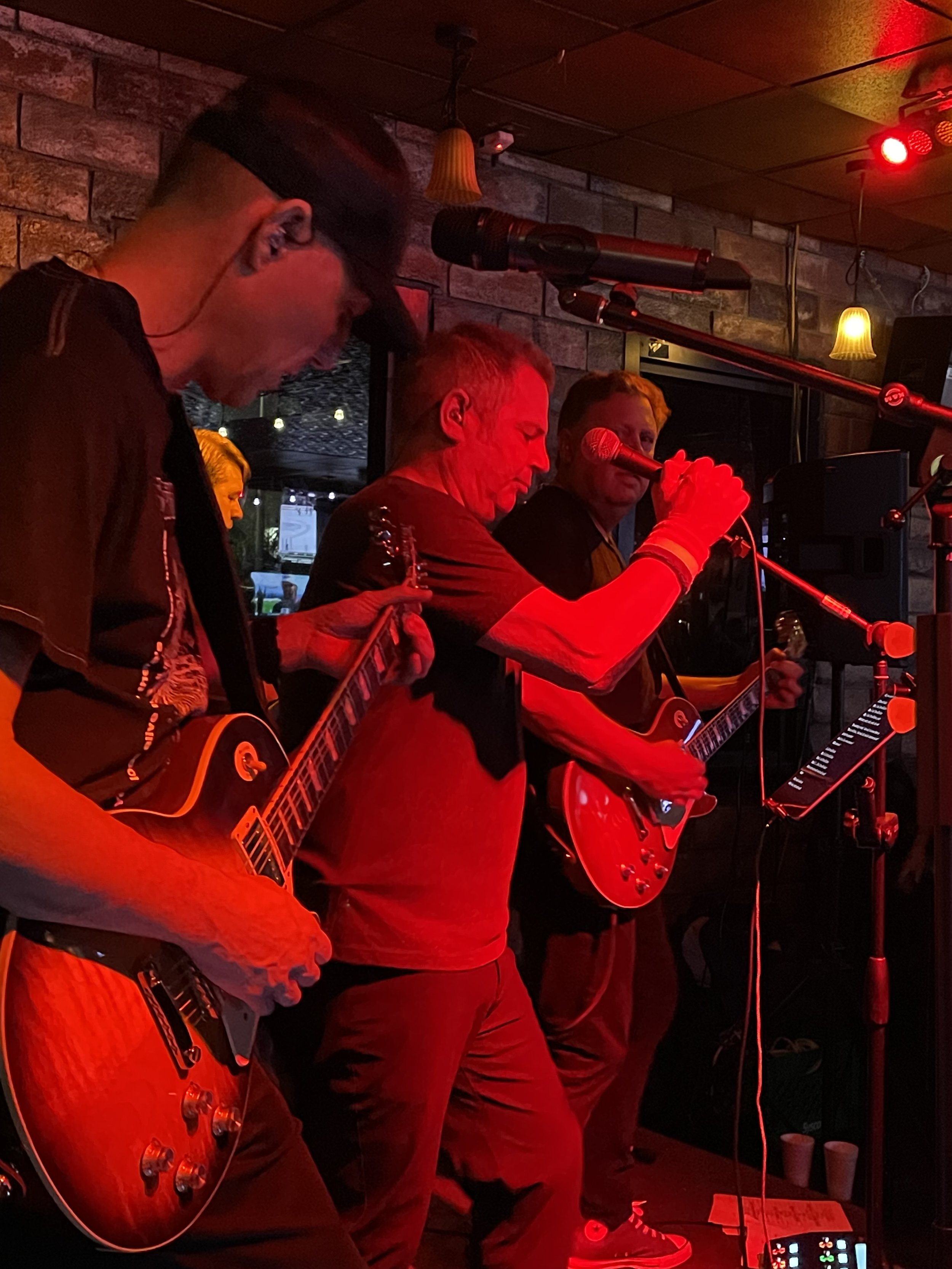 Three musicians playing electric guitars on stage in a dimly lit venue with red and yellow lighting and a brick wall background.