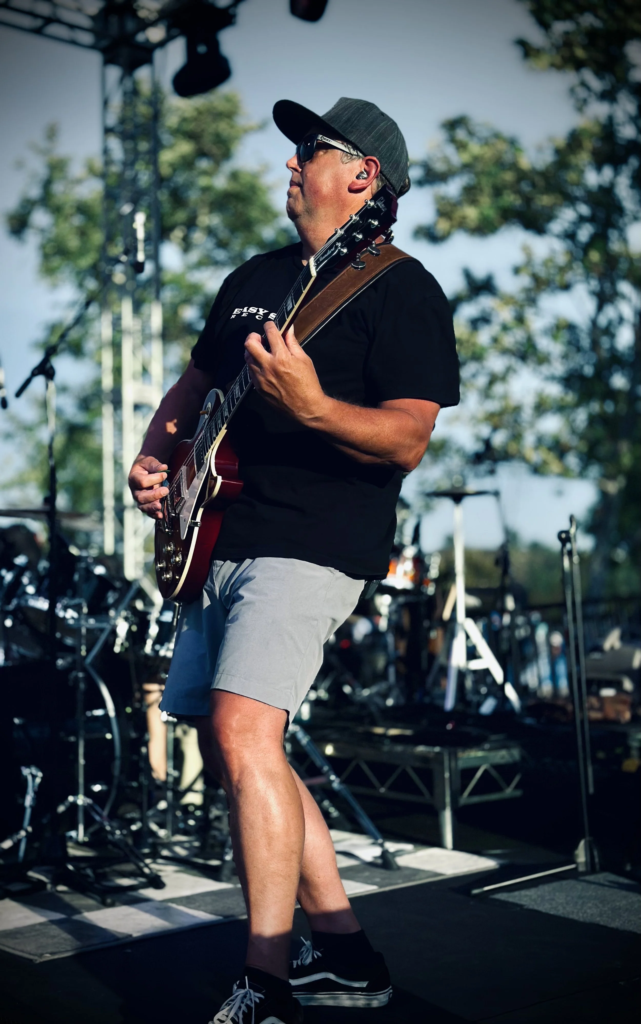 A man playing electric guitar on stage during an outdoor concert, wearing sunglasses, a black baseball cap, a black t-shirt, gray shorts, and sneakers, with trees and stage equipment in the background.