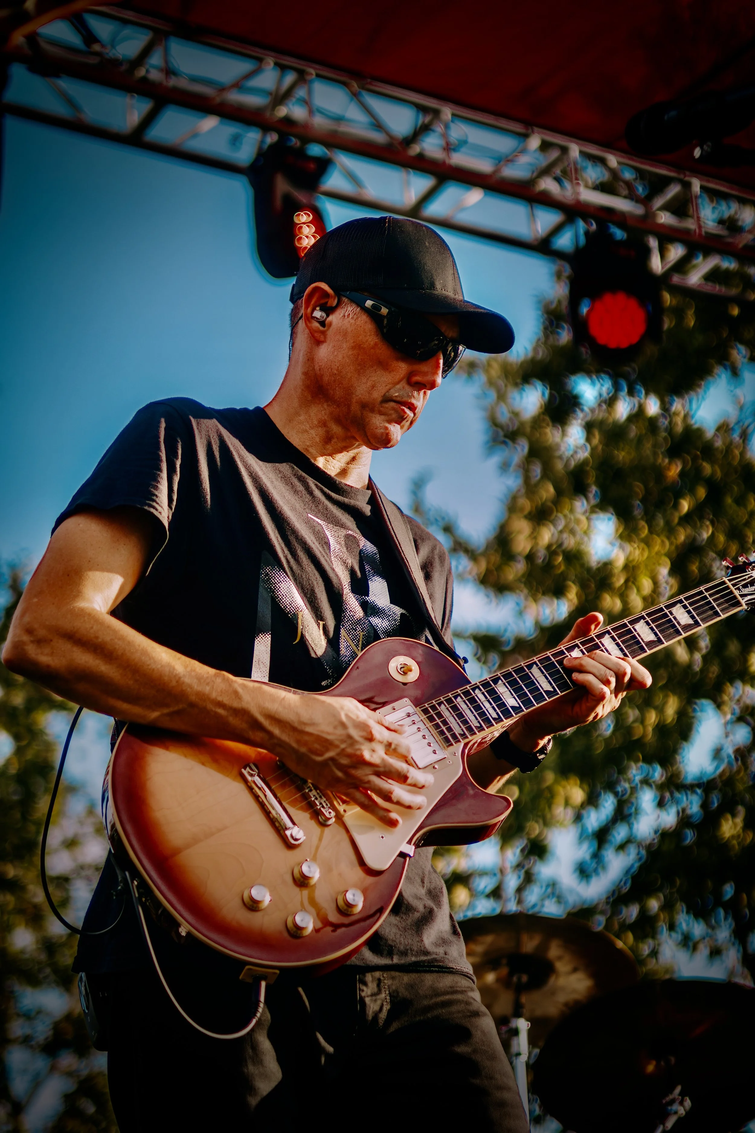 A man wearing a black cap, sunglasses, and a black t-shirt playing an electric guitar on an outdoor stage with trees in the background.
