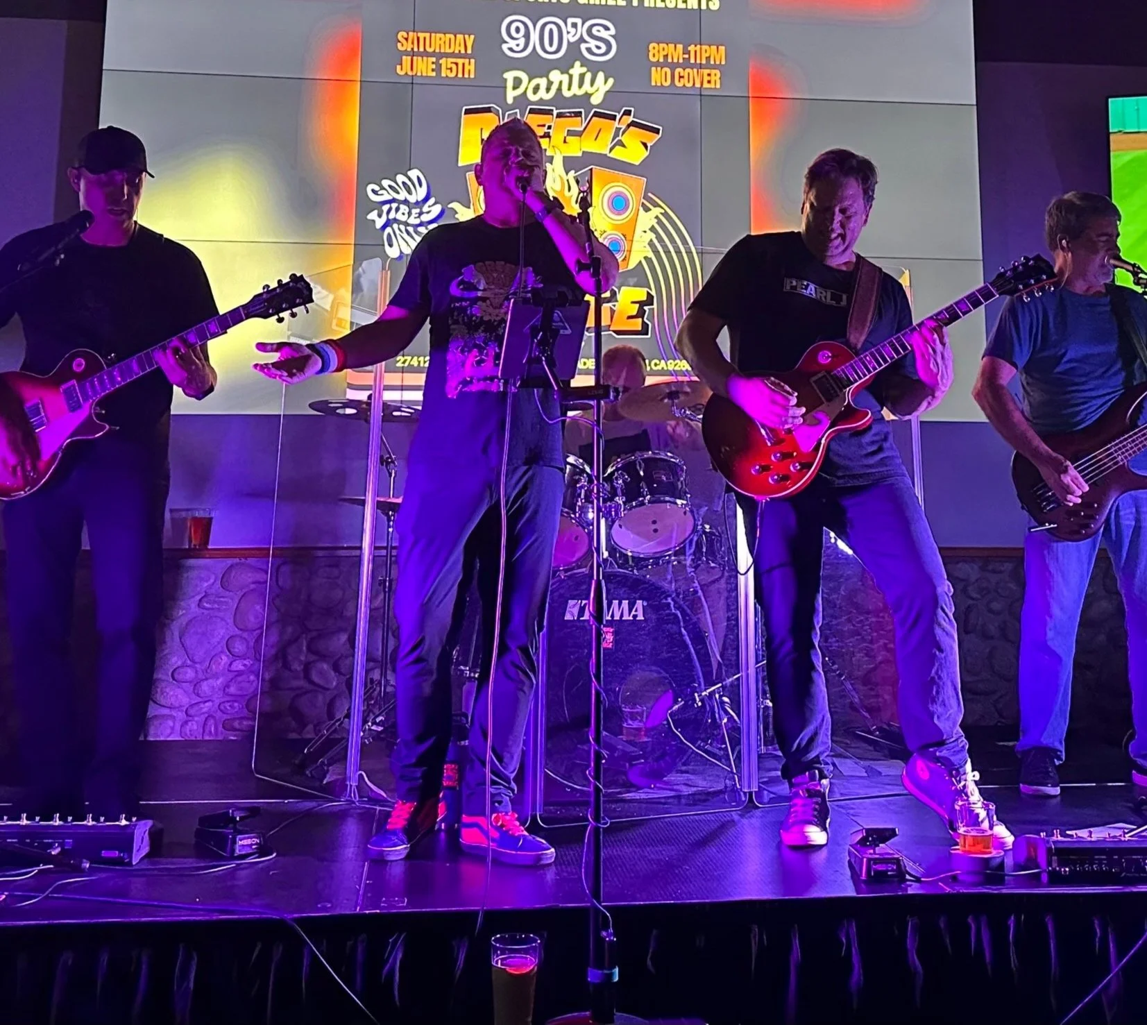 A band performing live on stage with four male musicians playing guitars and singing, colorful stage lighting, a drum set in the background, and a large digital display promoting a '90s themed party event.