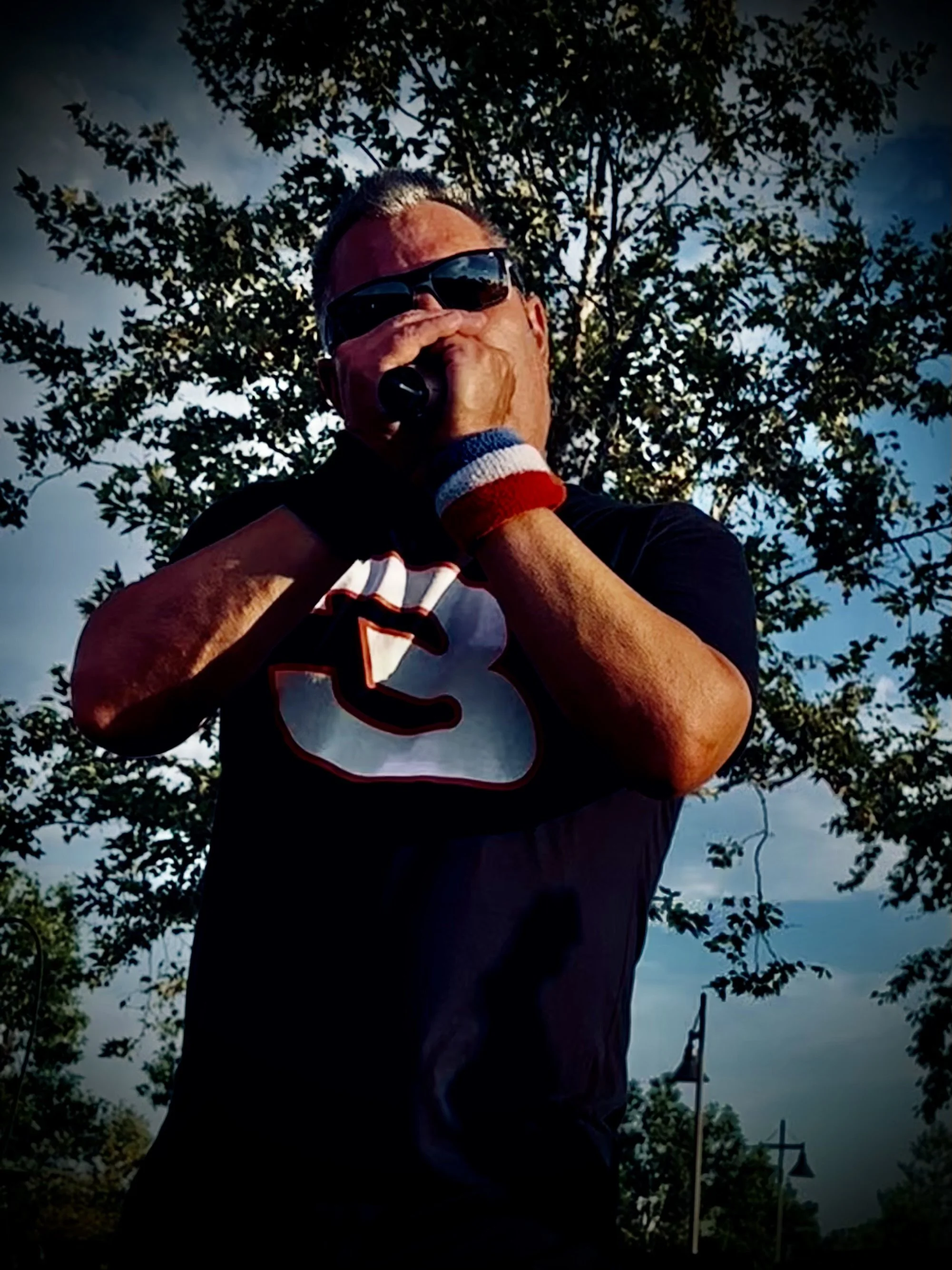 Man wearing sunglasses and a black T-shirt with a sports logo, holding a microphone, outdoors with trees and a cloudy sky in the background.