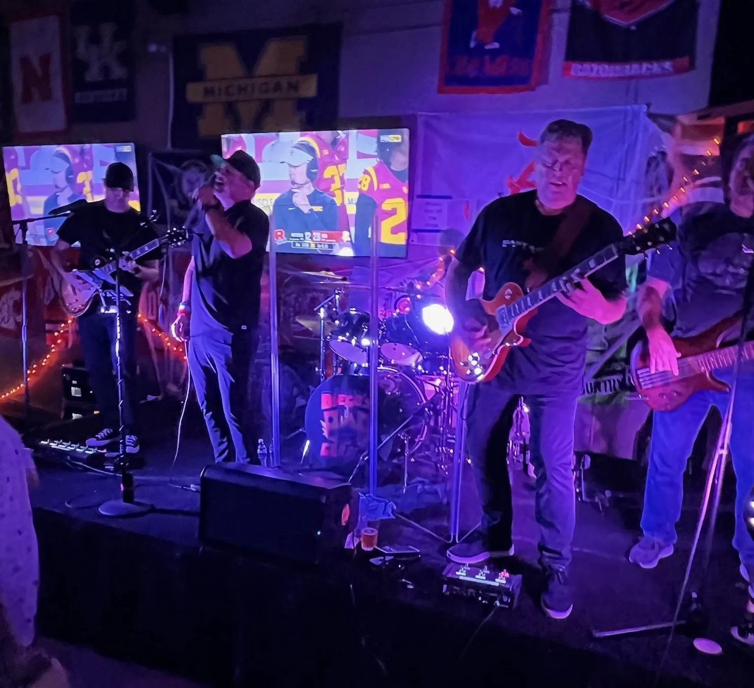 A band performing live music on a stage with purple lighting, featuring a guitarist, a singer, a drummer, and a bassist, in a bar or club decorated with sports banners and televisions.