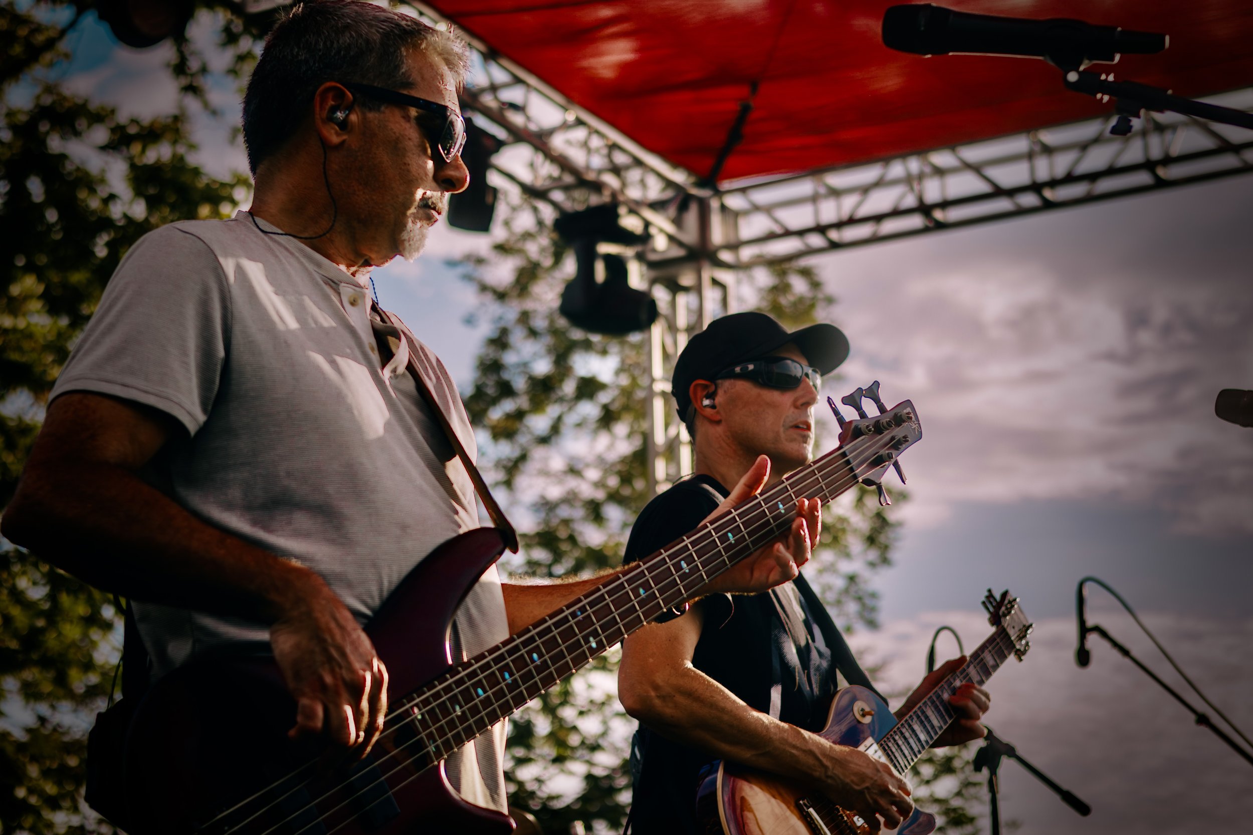Two male musicians playing guitars on an outdoor stage during sunset, with trees and cloudy sky in the background.