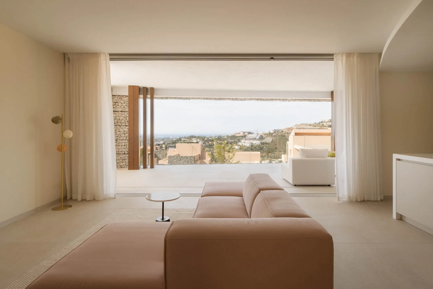 Minimalist living room with beige sofa, small round coffee table, white curtains, large window revealing a view of hills and buildings outside.