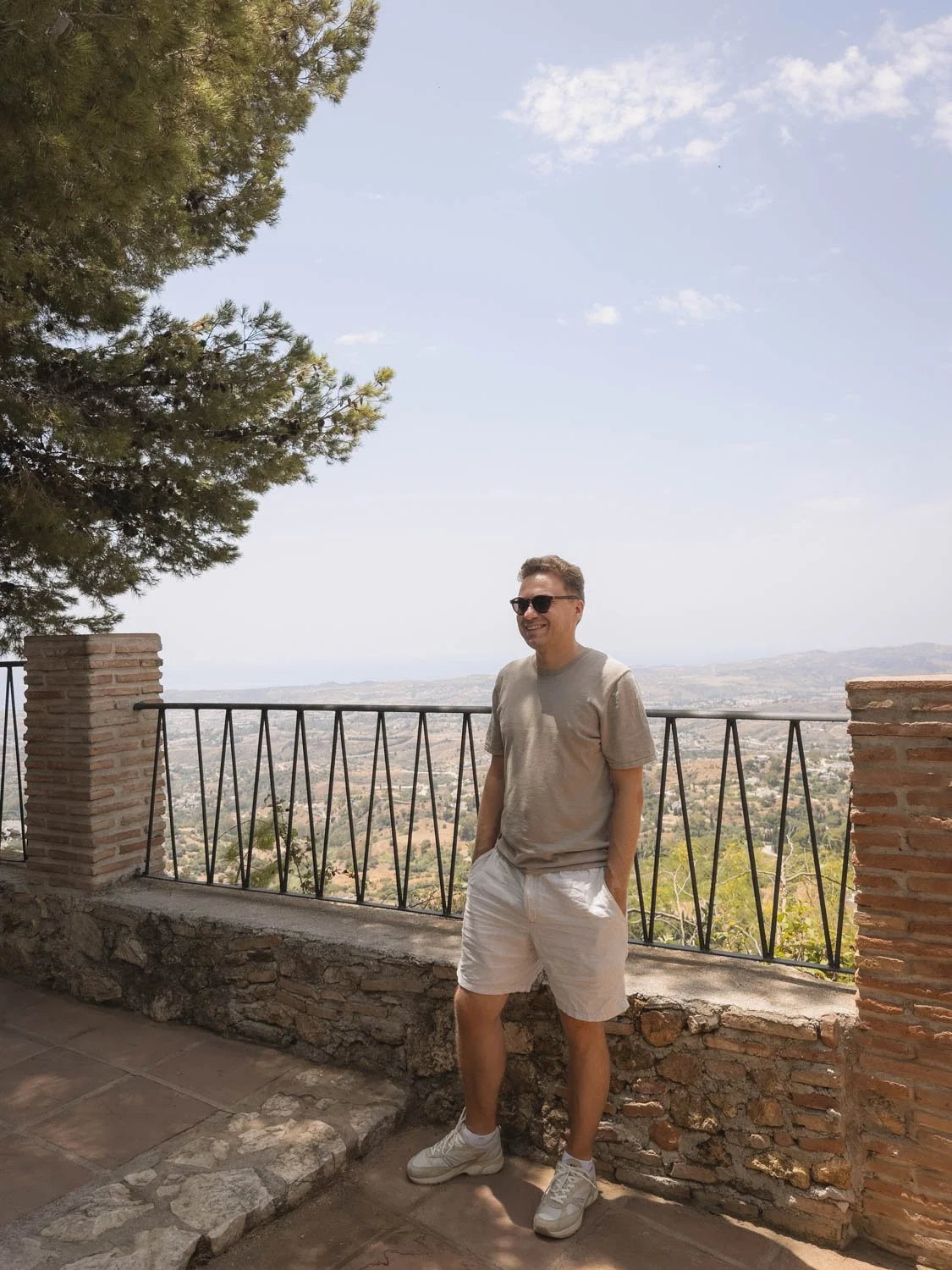 A man wearing sunglasses, a beige t-shirt, and white shorts standing by a brick and metal railing overlooking a landscape with trees and hills under a partly cloudy sky.