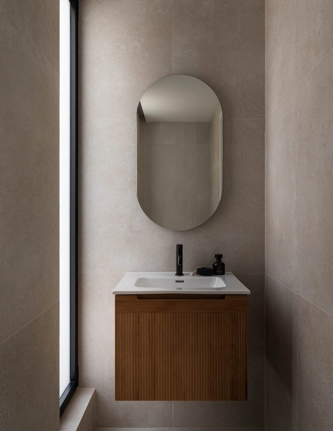 Modern bathroom sink with a wooden cabinet, an oval mirror, black faucet, and black soap dispenser, with a window to the left.