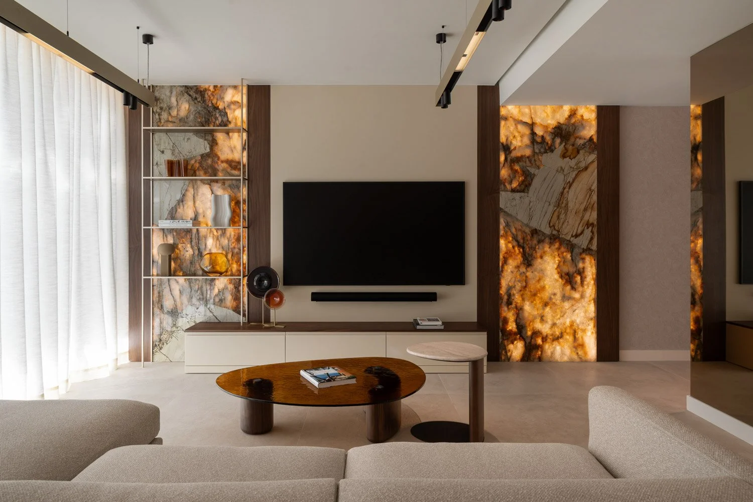 Modern living room with a large flat-screen TV on the wall, decorative rocky backlit panels, a beige sofa, a wooden coffee table with magazines, and shelves with decorative items.