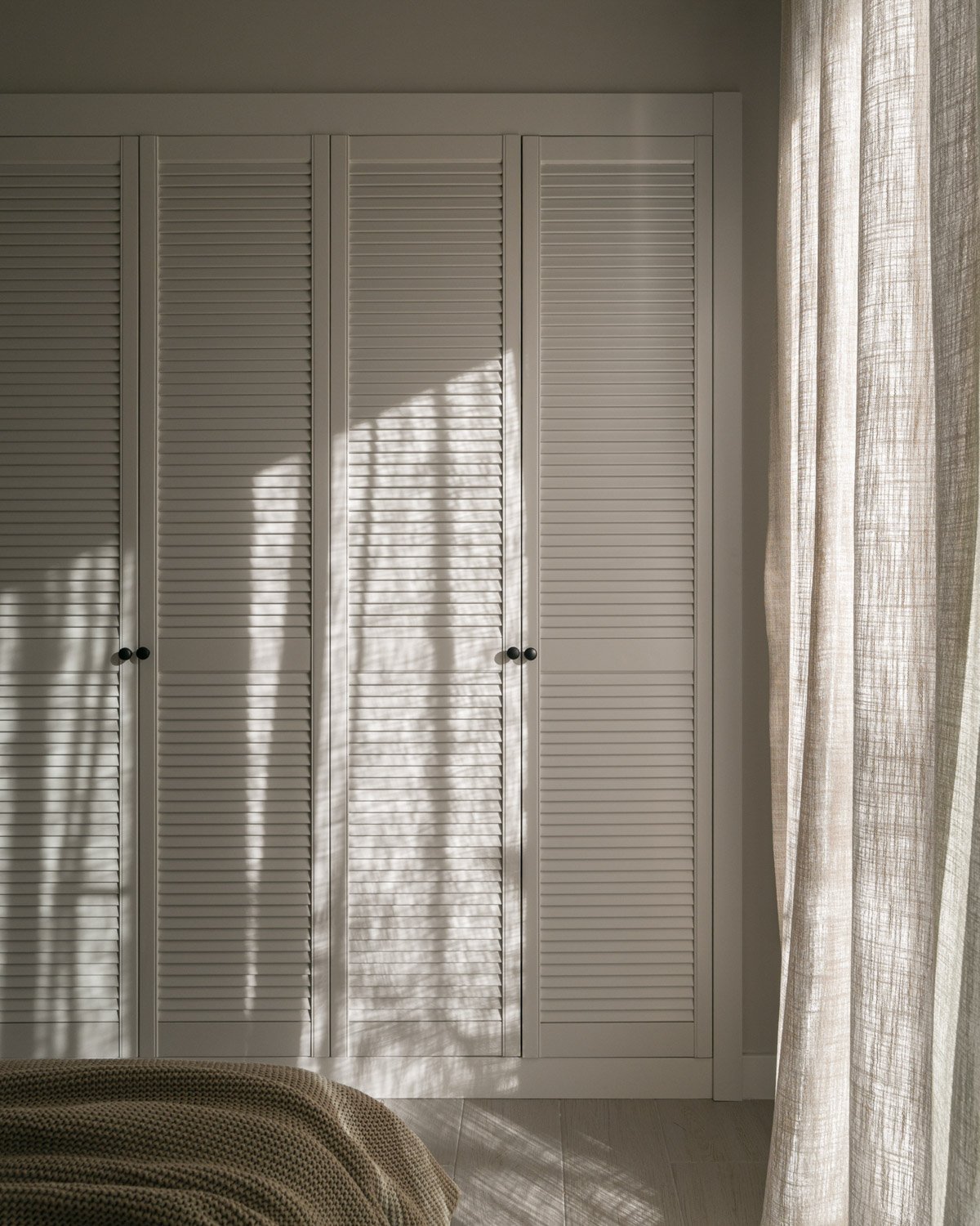 Sunlight casting shadows on white louvered wardrobe doors and linen curtains.