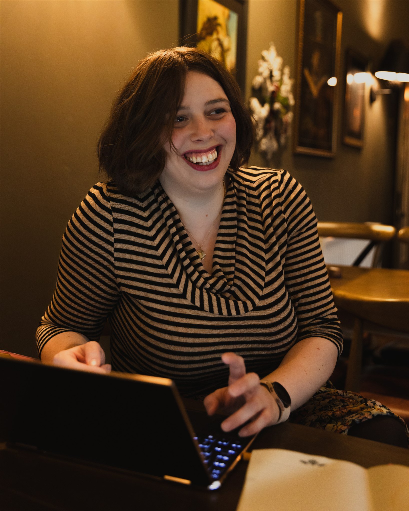 A young woman with short brown hair, smiling and typing at a laptop inside a cafe.