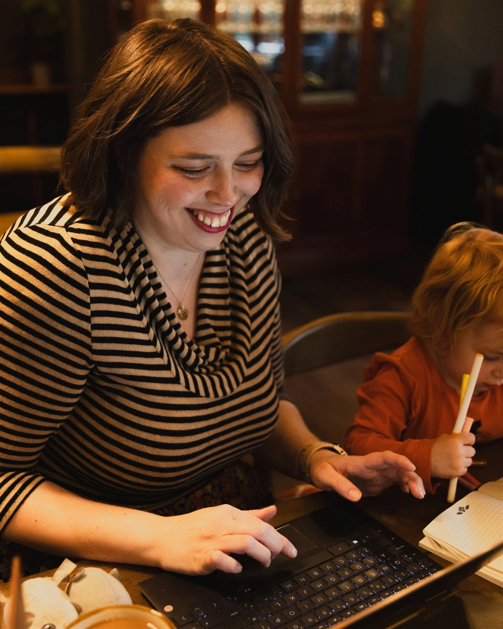 A woman with short, wavy brown hair smiling and using a laptop in a cafe.