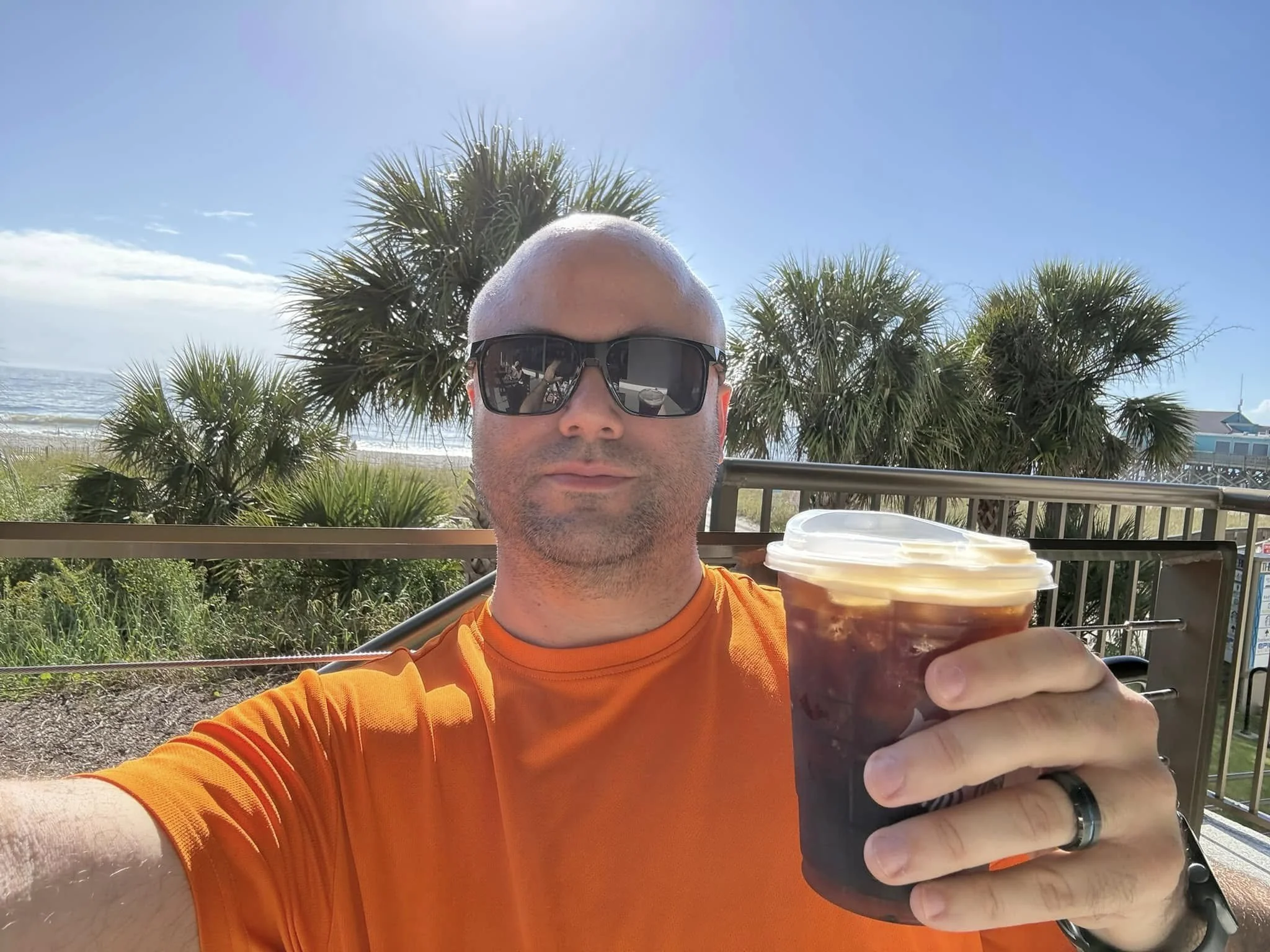 A bald man wearing sunglasses and an orange shirt holding a cup of iced coffee at an outdoor location near the beach, with palm trees and a blue sky in the background.