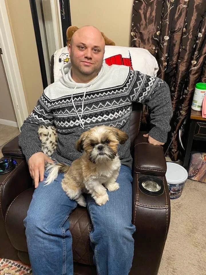 A man with a shaved head sitting in a brown recliner, wearing a patterned holiday sweater, with a small fluffy dog sitting on his lap. The environment appears cozy with curtains and household items in the background.