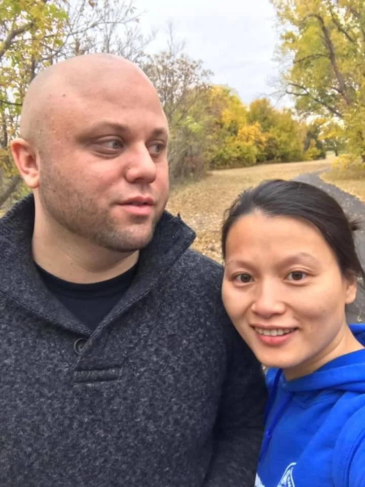 A man and woman taking a selfie outdoors on a path during autumn, with trees showing fall foliage in the background.