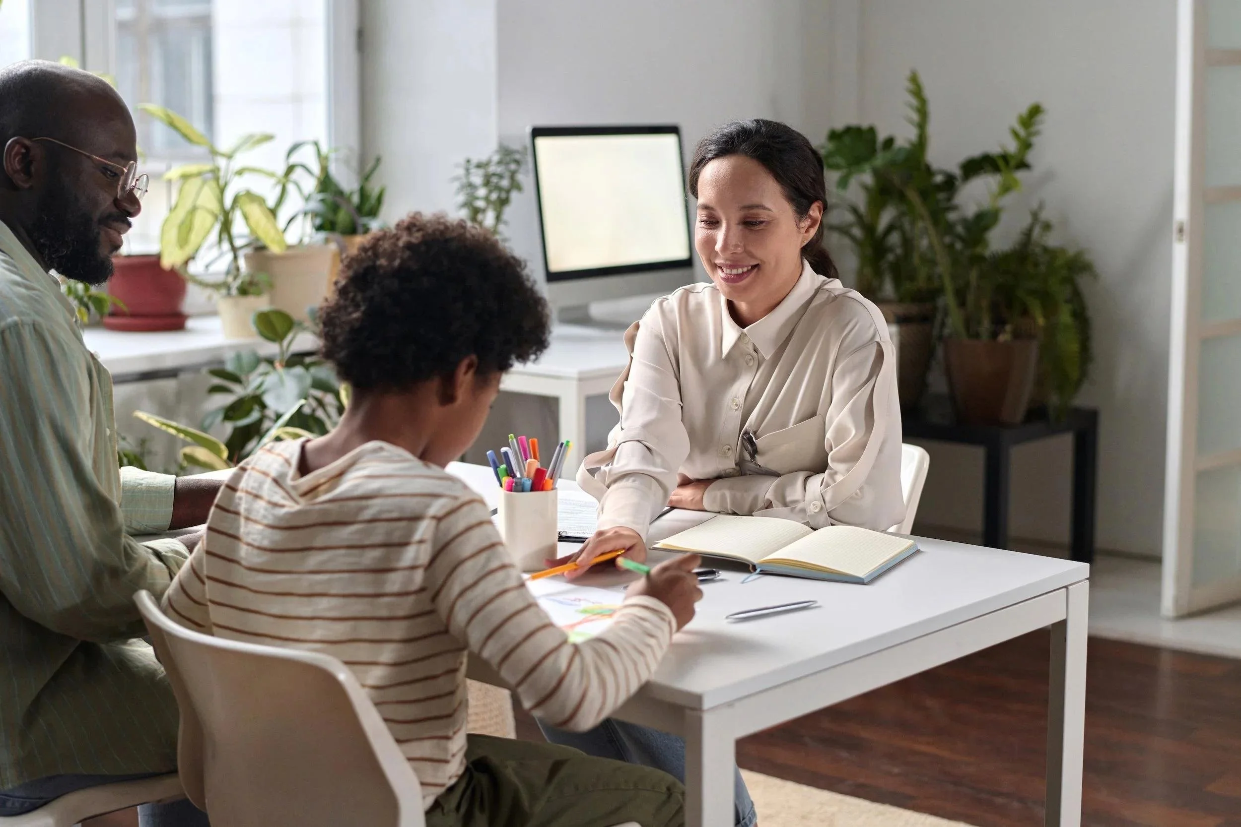 Child psychologist guiding a child through a drawing activity with a parent seated beside them in a bright, welcoming office.