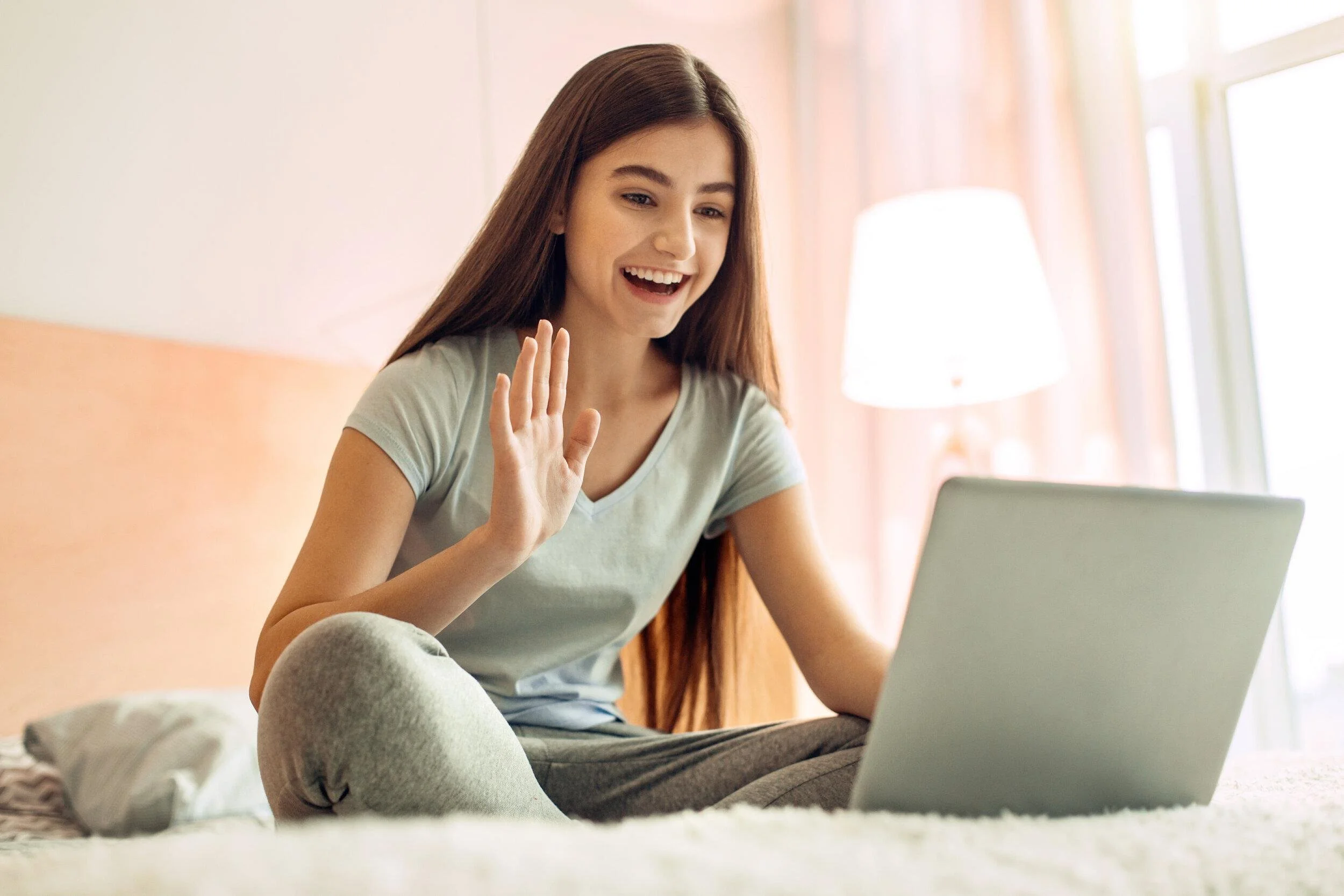 Teenager smiling and waving during a laptop video call in her bedroom, illustrating virtual emotional regulation therapy.