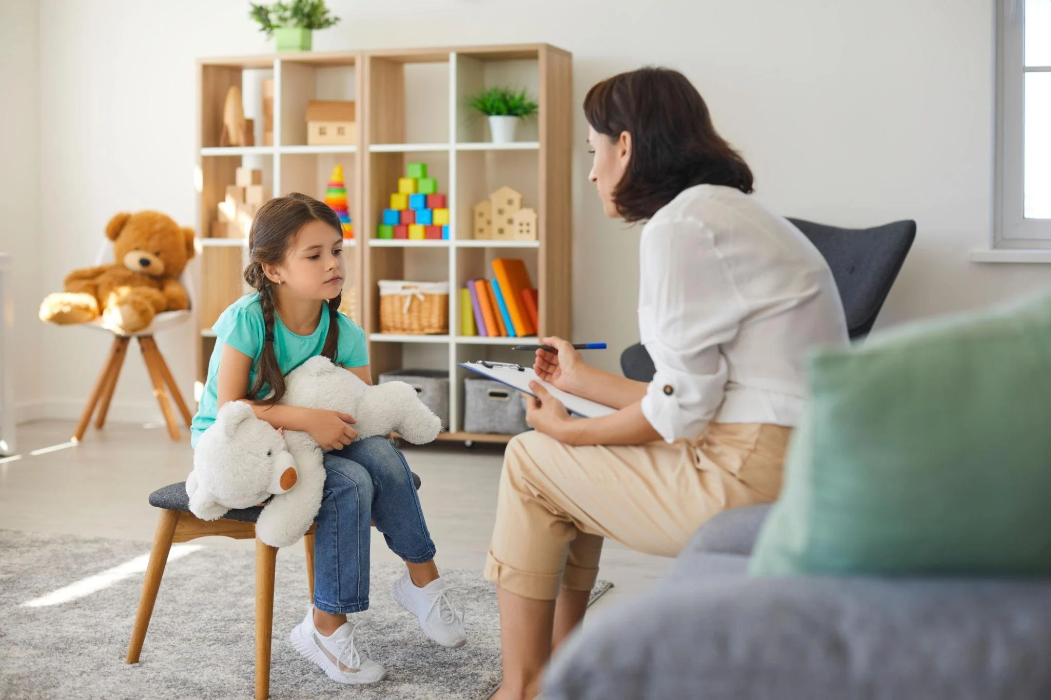 Young girl holding a stuffed animal while talking with a counselor in a bright, child-friendly therapy room.