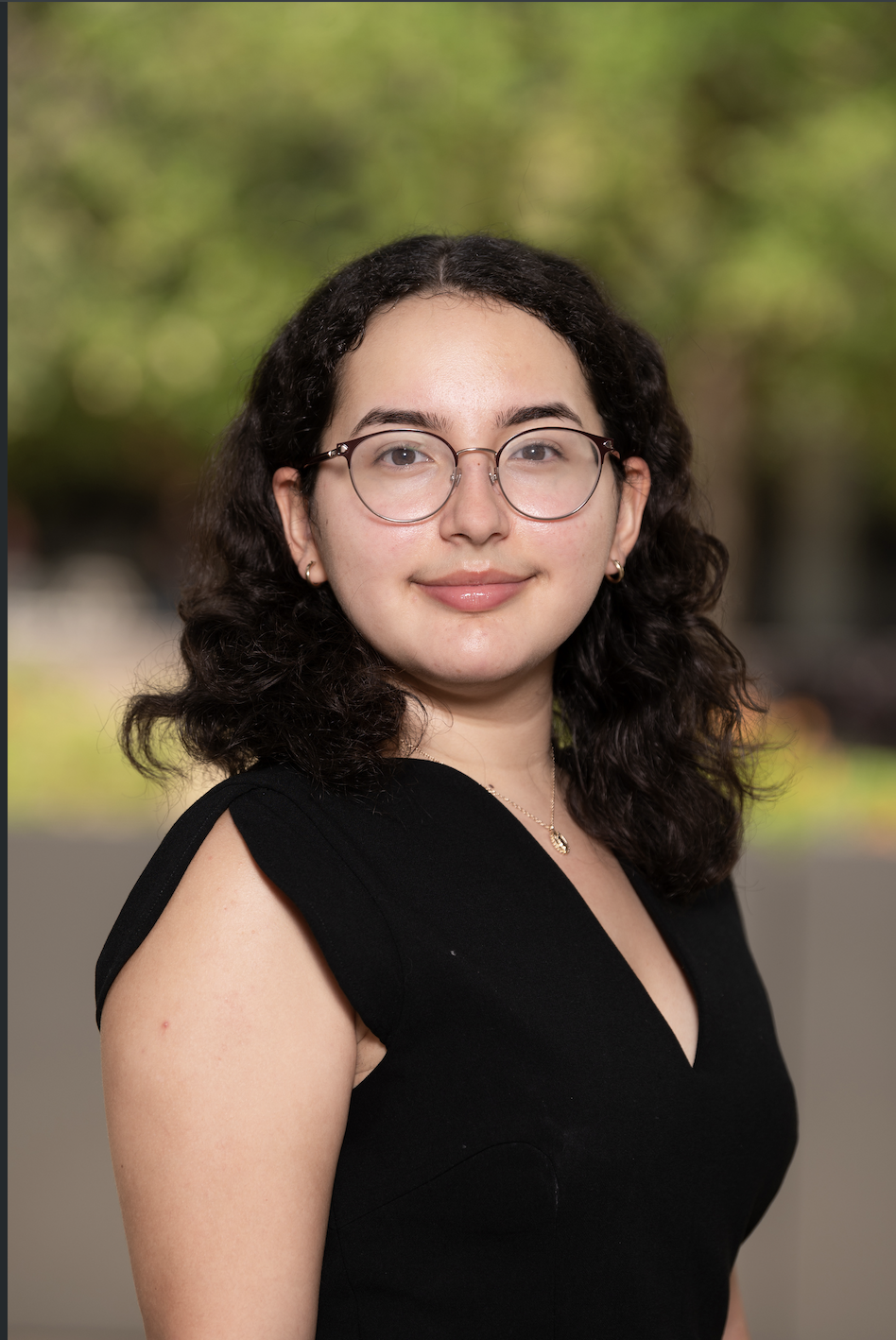 A portrait of a young woman with shoulder-length curly dark hair, wearing glasses, a black sleeveless dress, a gold necklace, and earrings, standing outdoors with blurred green foliage in the background.