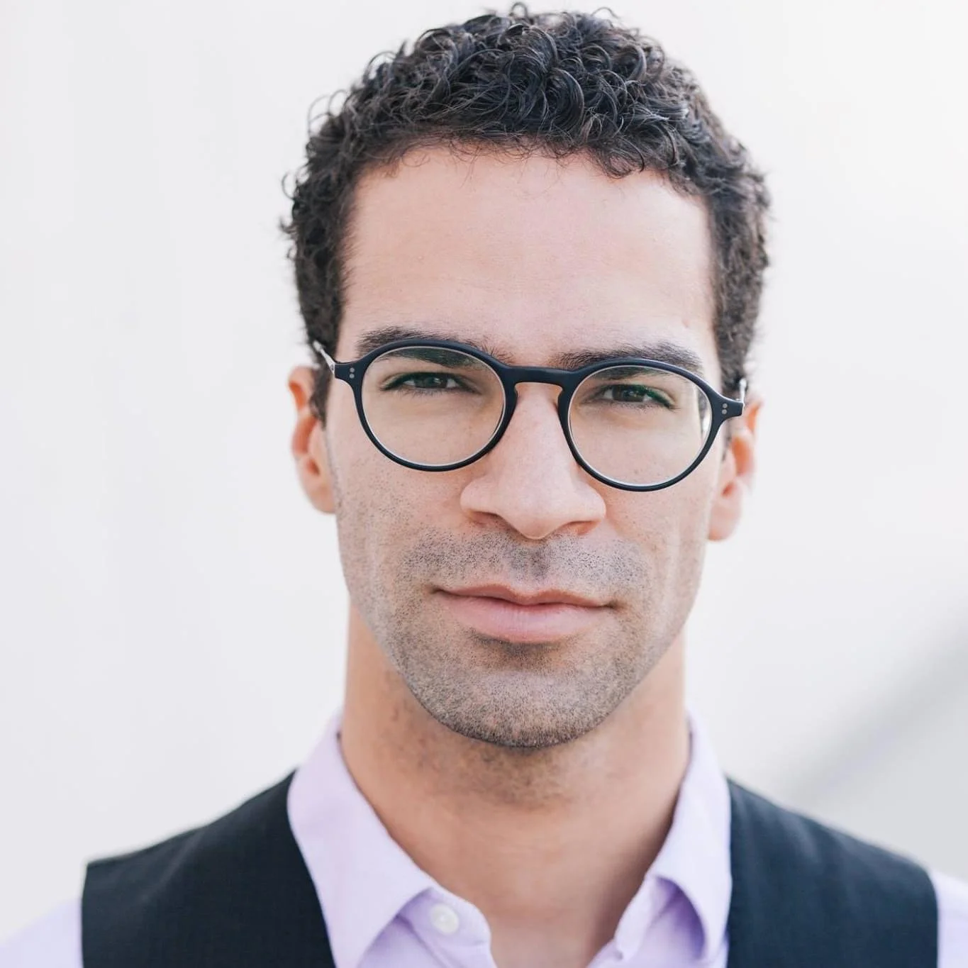 A young man with short dark curly hair, wearing black round glasses, a light purple dress shirt, and a black vest against a plain, light background.