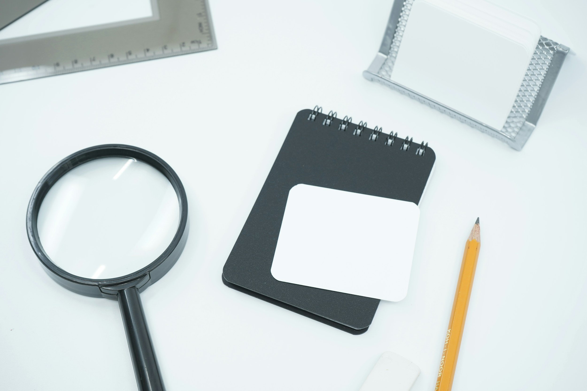 A flat lay of office supplies on a white surface, including a magnifying glass, rulers, a black spiral notepad, a small blank white notepad, and a yellow pencil.