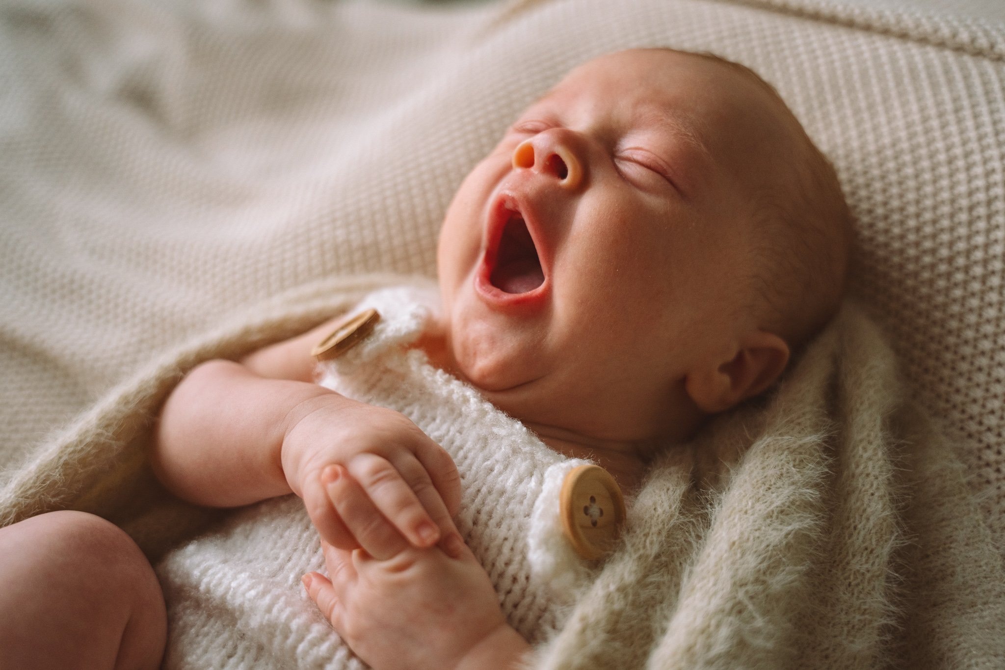 A baby yawning, lying on a beige textured blanket, dressed in a knit sweater with buttons.