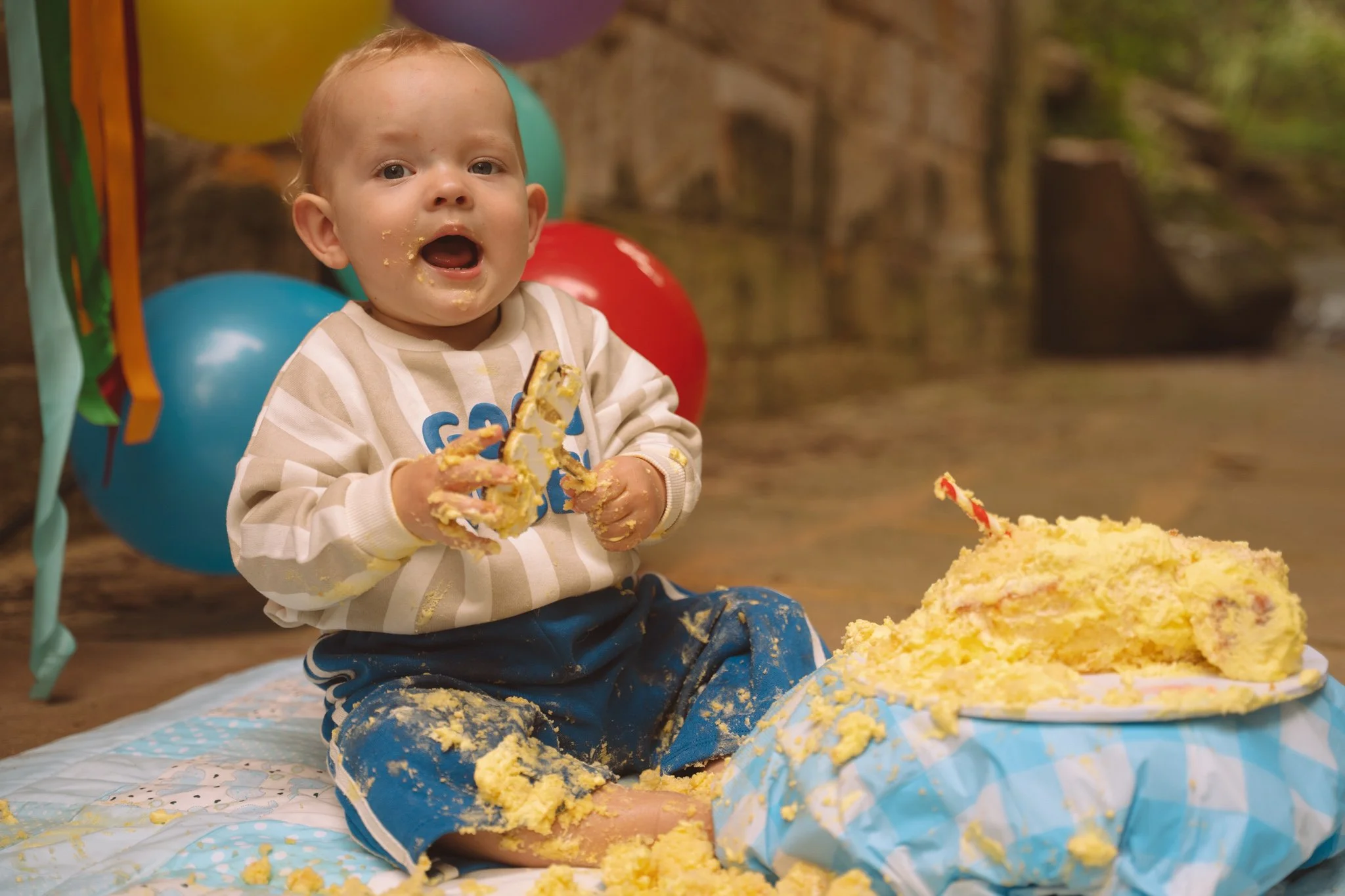 A young child with short hair and wearing a striped shirt sitting on a quilted blanket with cake and yellow frosting all over their hands and face. The child is surrounded by colorful balloons and appears to be celebrating a birthday.