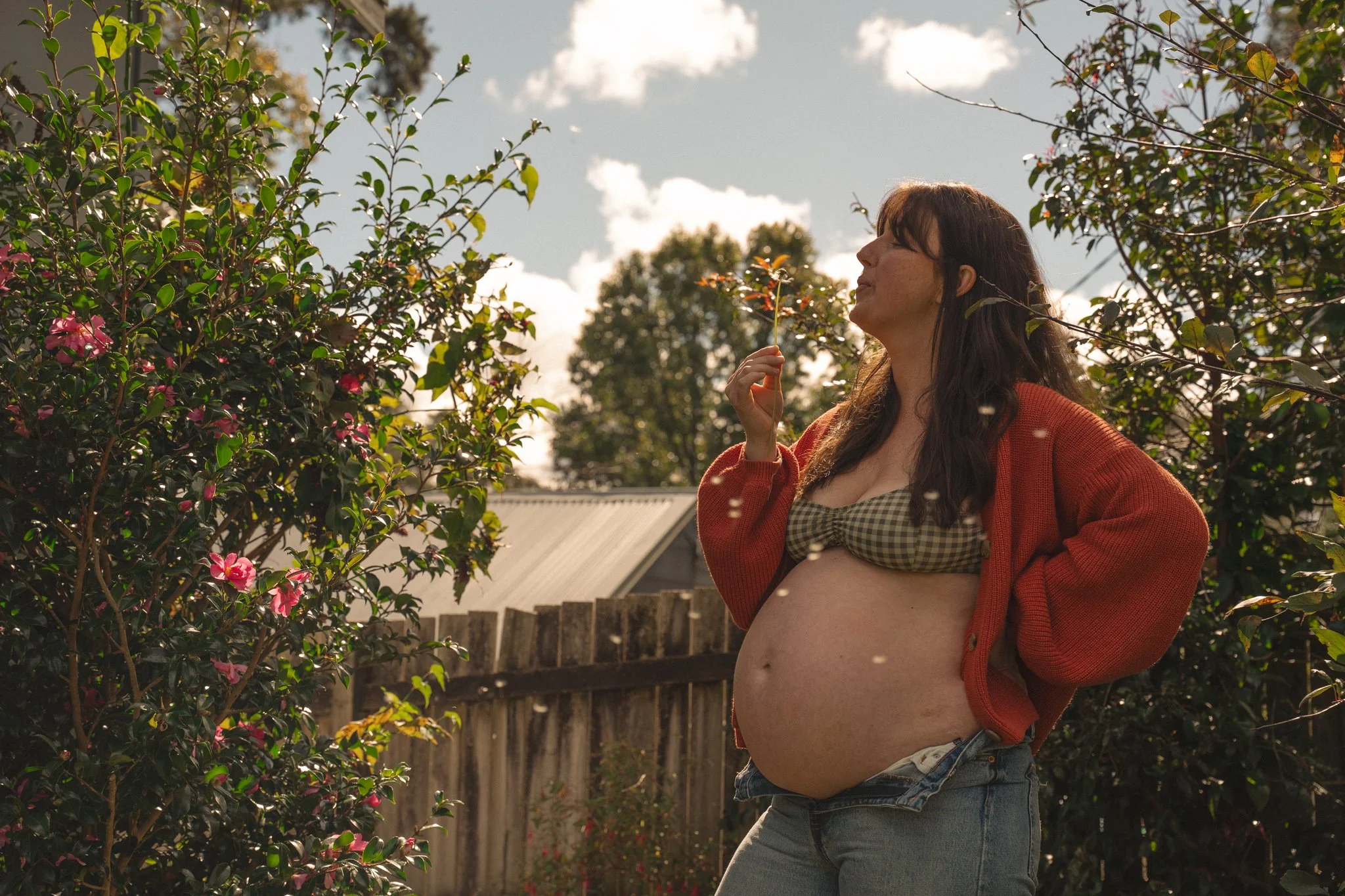 A pregnant woman with long dark hair is standing outdoors in a backyard, wearing a checkered top and unbuttoned red cardigan. She is holding a flower close to her nose with her eyes closed, surrounded by green bushes with pink flowers and trees under a partly cloudy sky.