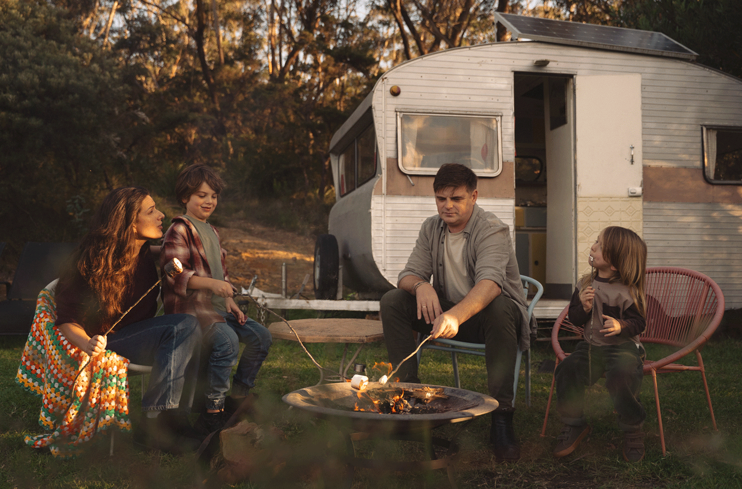 Family camping outdoors near a caravan, roasting marshmallows over a fire pit, with trees in the background during sunset.