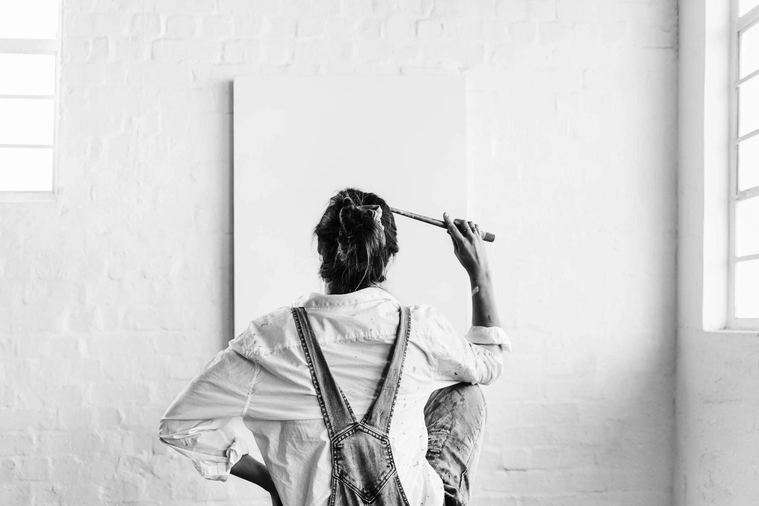 Artist sitting on floor in front of blank canvas, holding paintbrush to head, in bright studio with brick wall and large window.