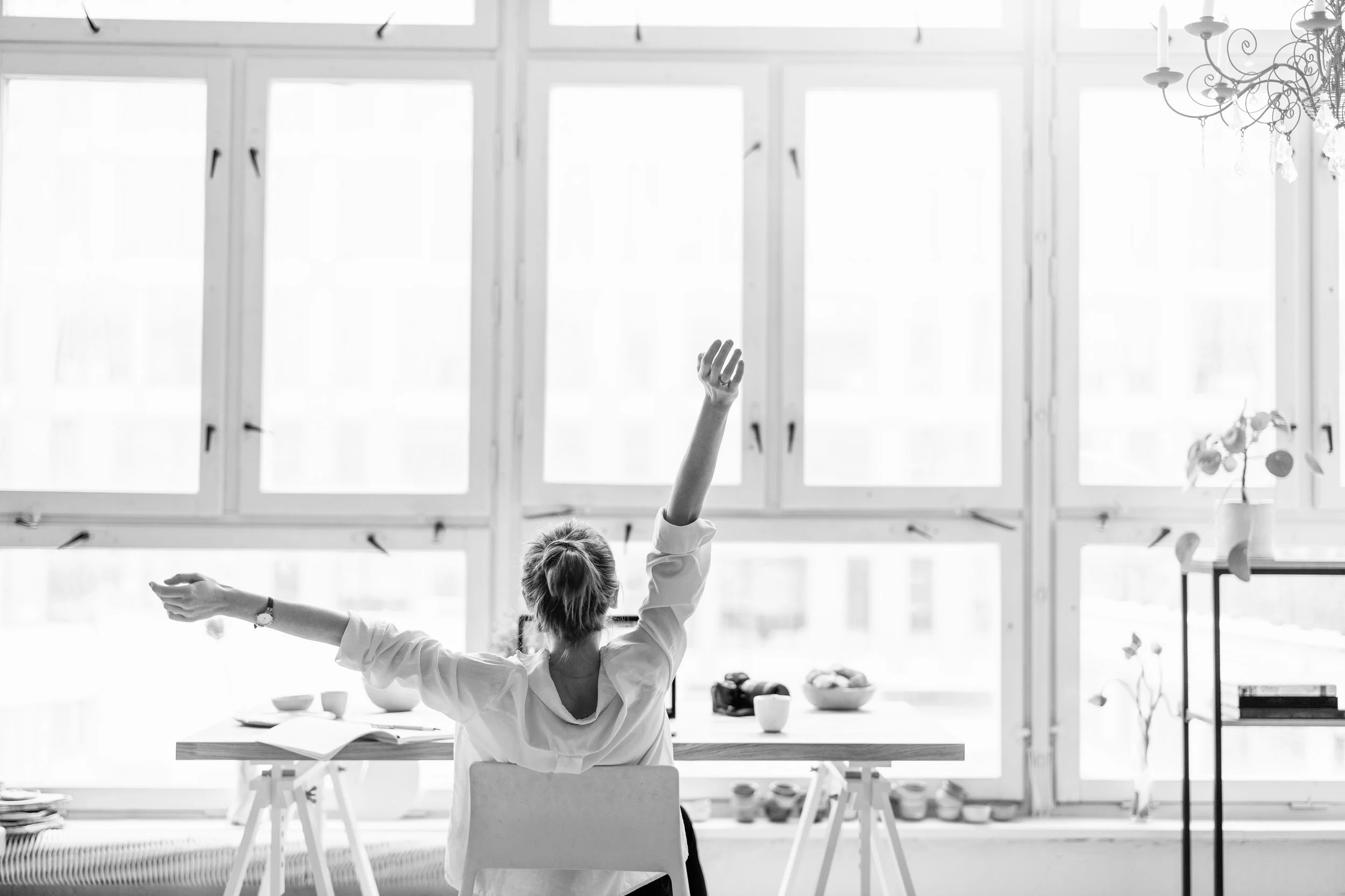 Black & white portrait of a woman sitting at a desk, with her arms over her head, looking out a window