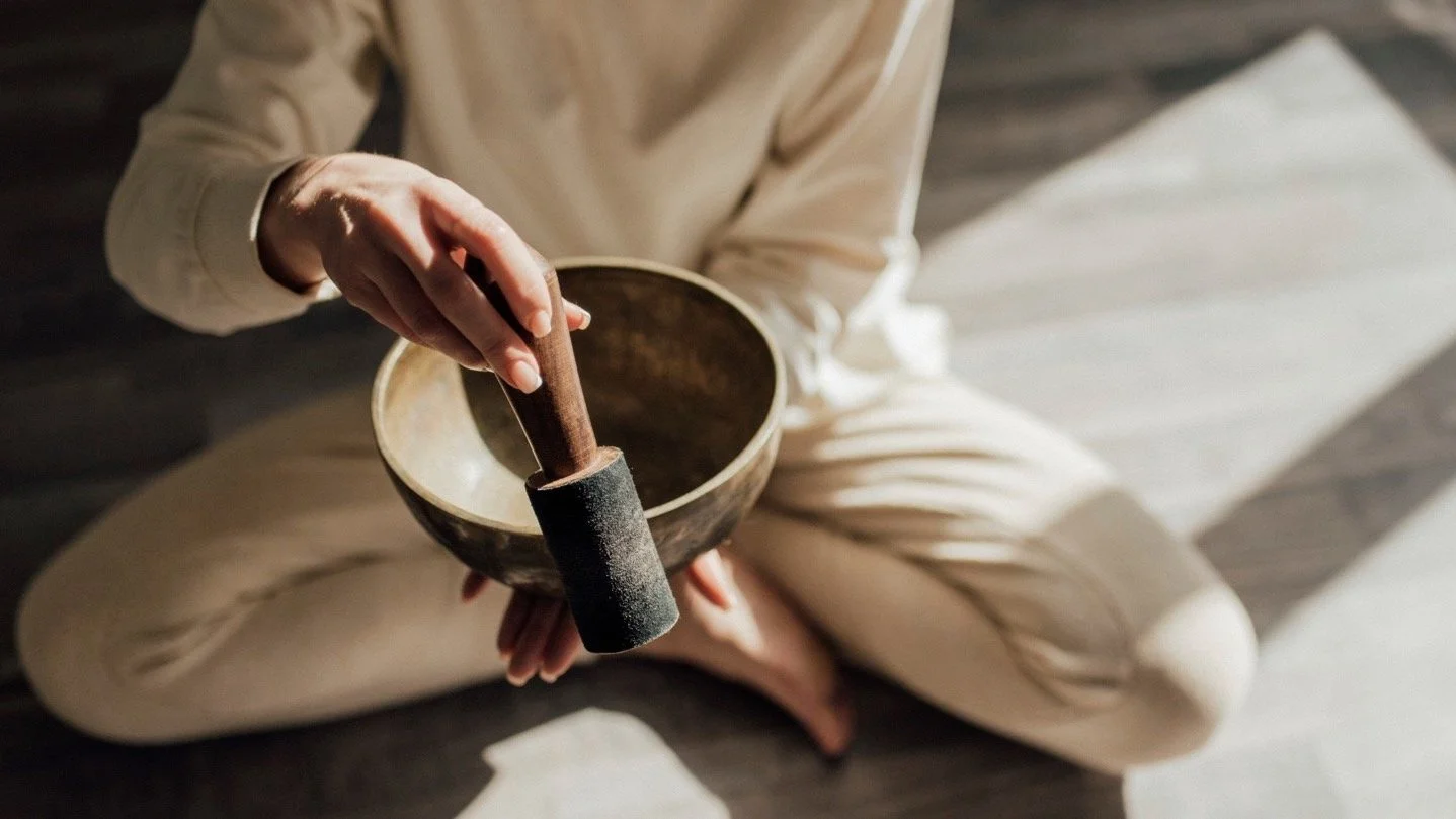 Person sitting cross-legged, holding a singing bowl and mallet, on a wooden floor with a striped rug nearby.