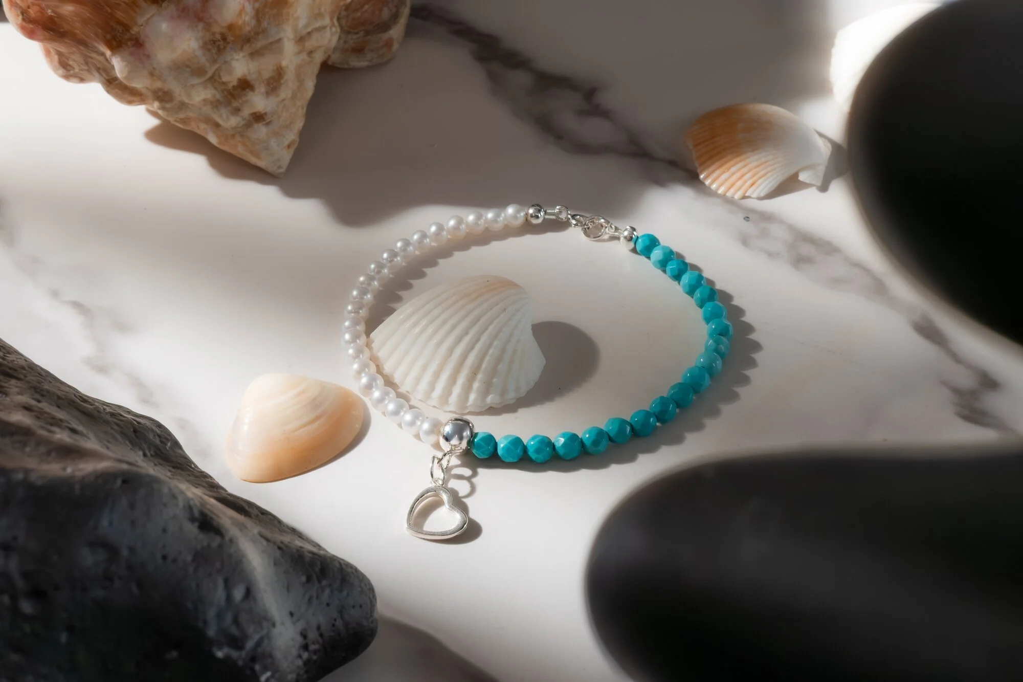 A beaded bracelet with blue and white beads and a silver heart-shaped charm, surrounded by seashells and rocks on a marble surface.