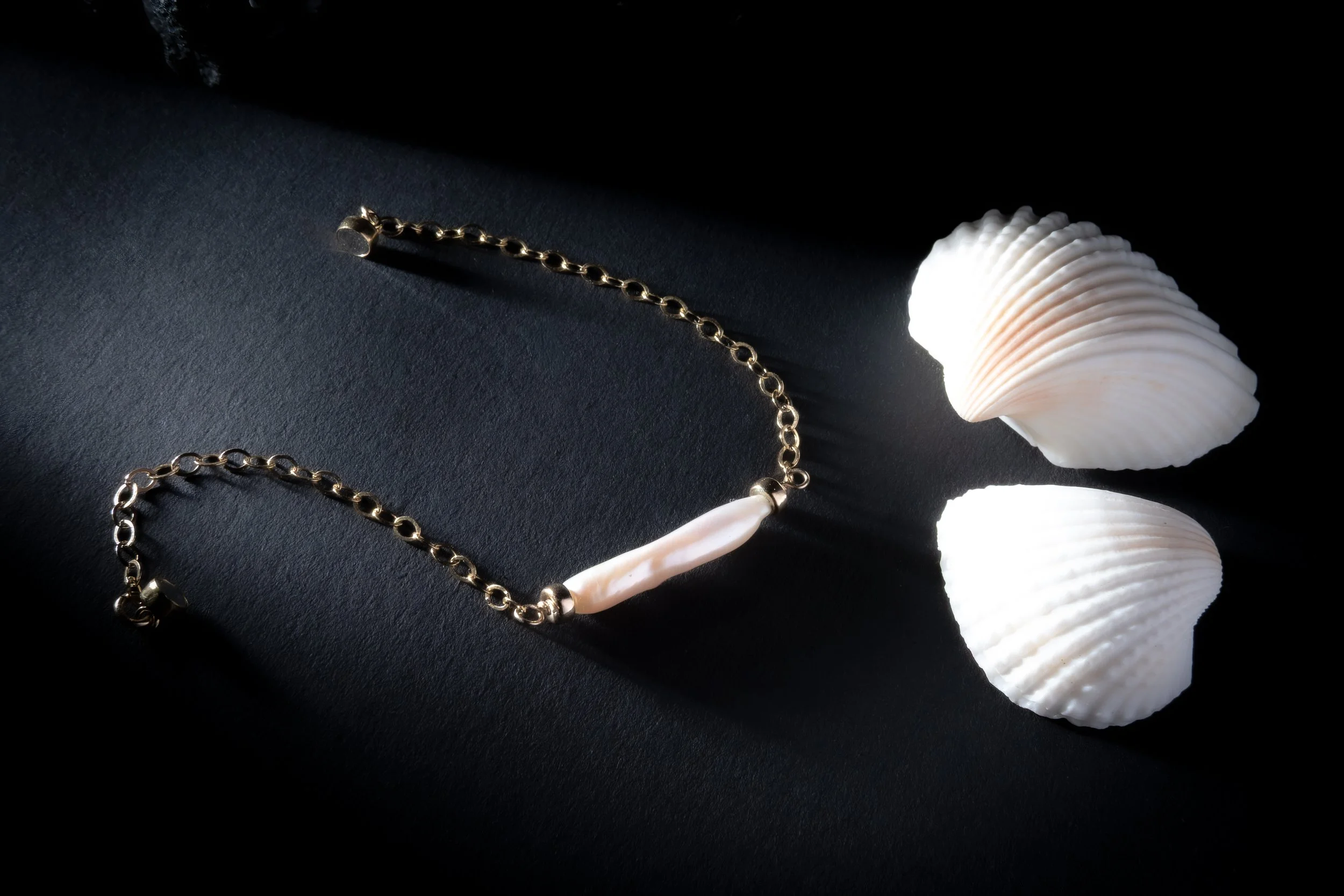 A gold chain necklace with a white pearl pendant placed on a dark surface next to two white seashells in sunlight.