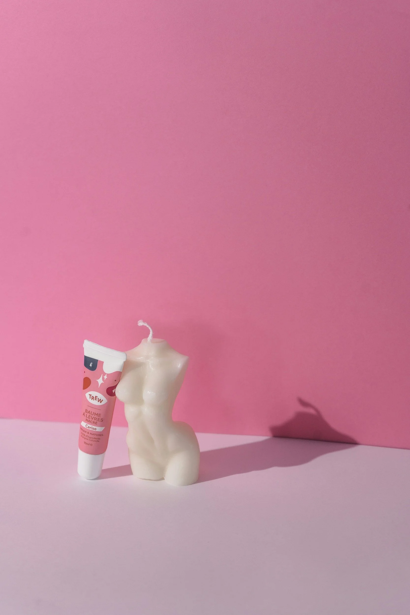 A pink background with a white candle shaped like a female torso and a small tube of Trew lip balm on a pink surface, casting shadows.