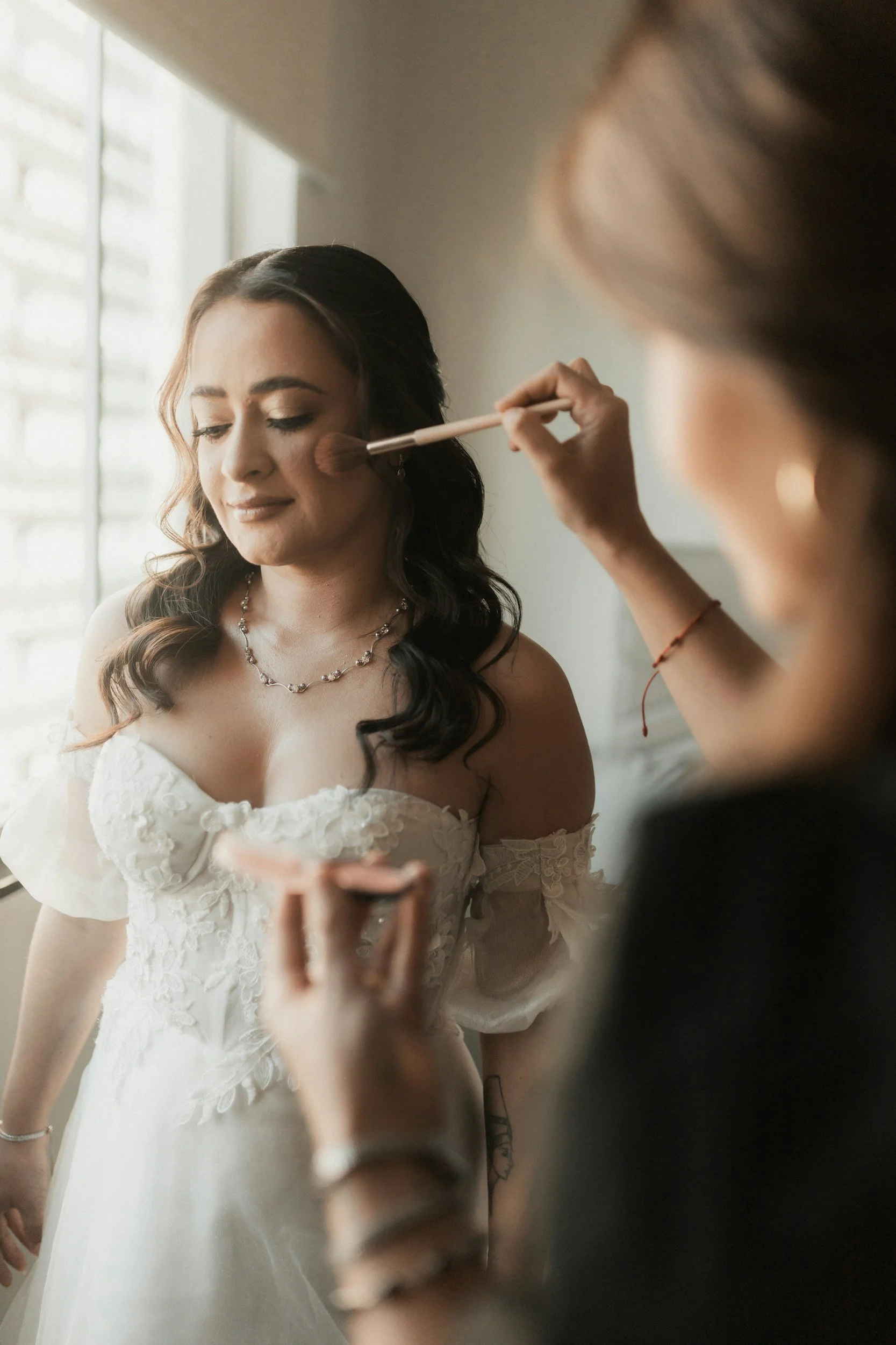 A woman in a wedding dress has makeup applied by a makeup artist using a brush near her cheek.