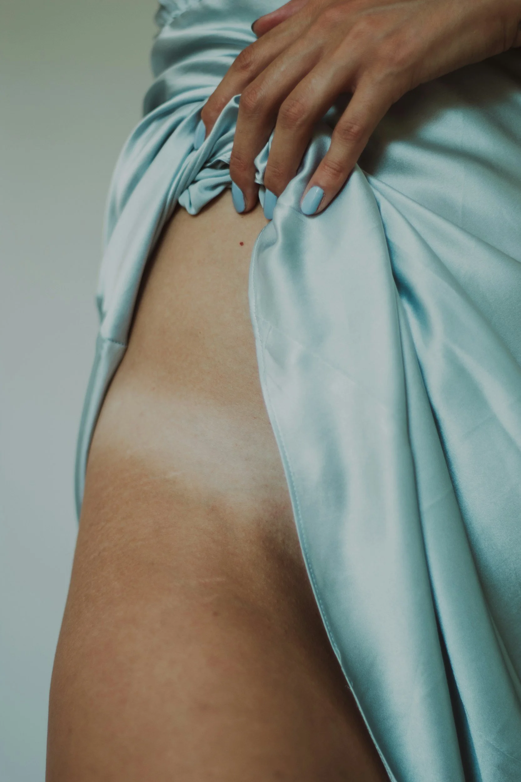 A person's hand lifting a light blue satin fabric, revealing thigh and a small mole on the skin.