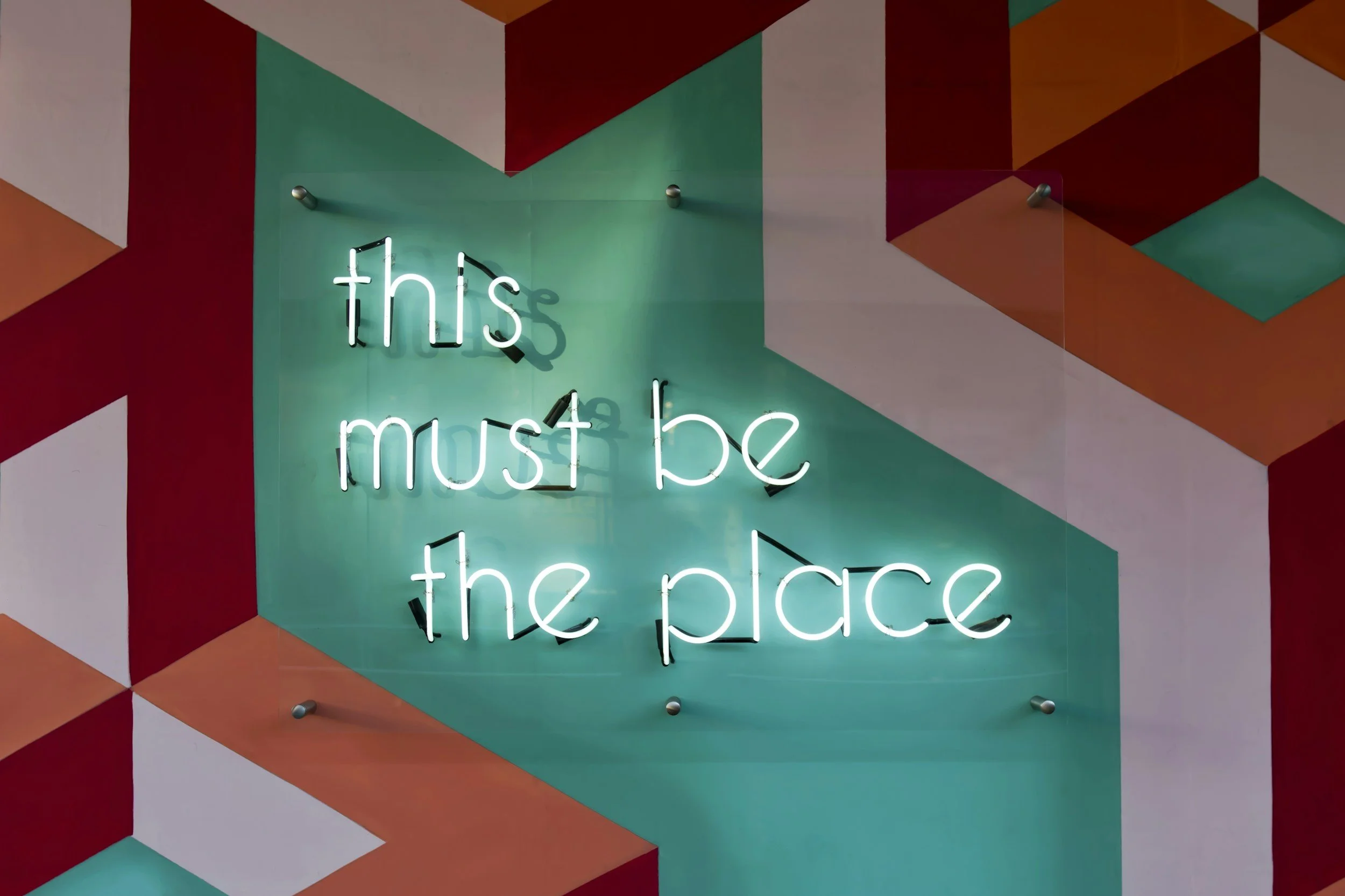 Neon sign with the phrase 'this must be the place' against a colorful geometric wall background.