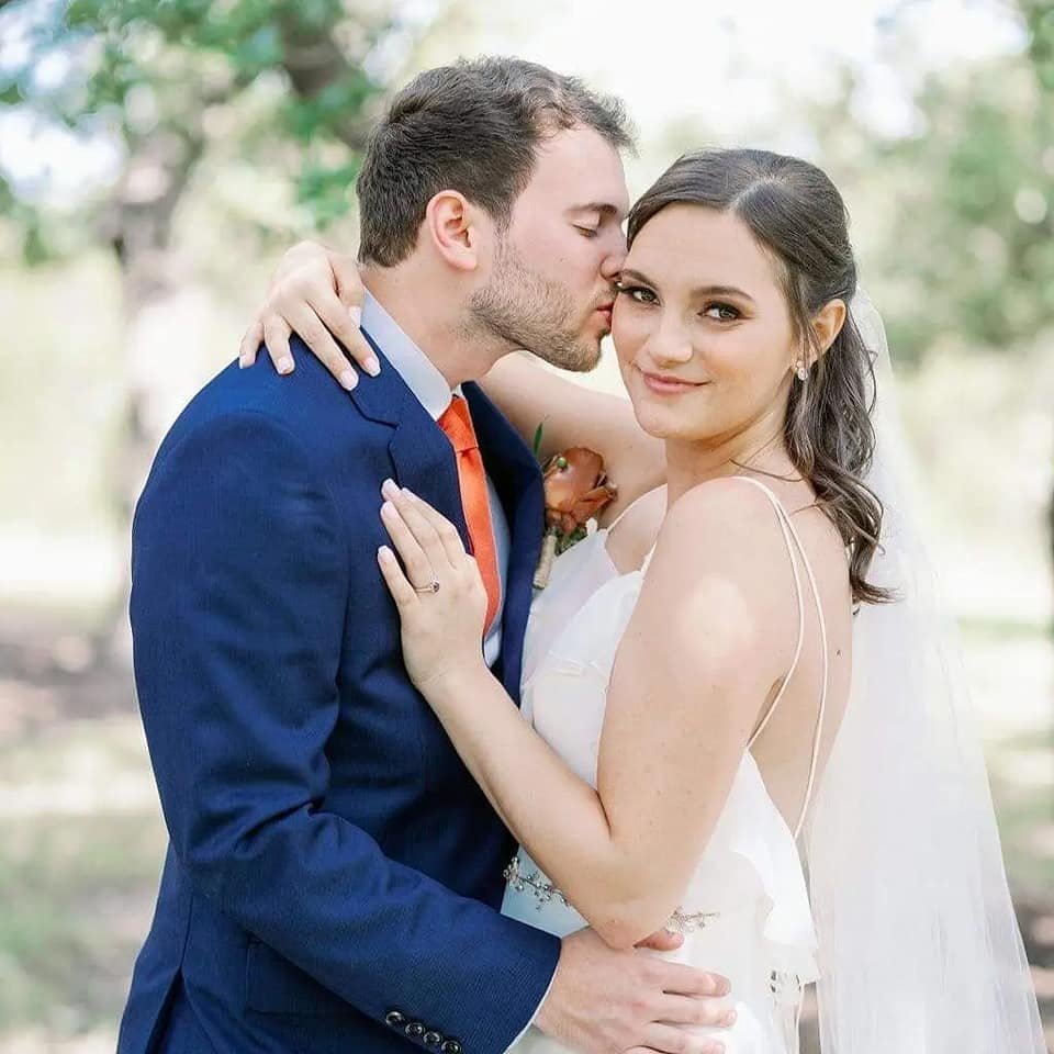 A bride and groom embrace outdoors, with the groom kissing the bride's temple while she smiles at the camera.