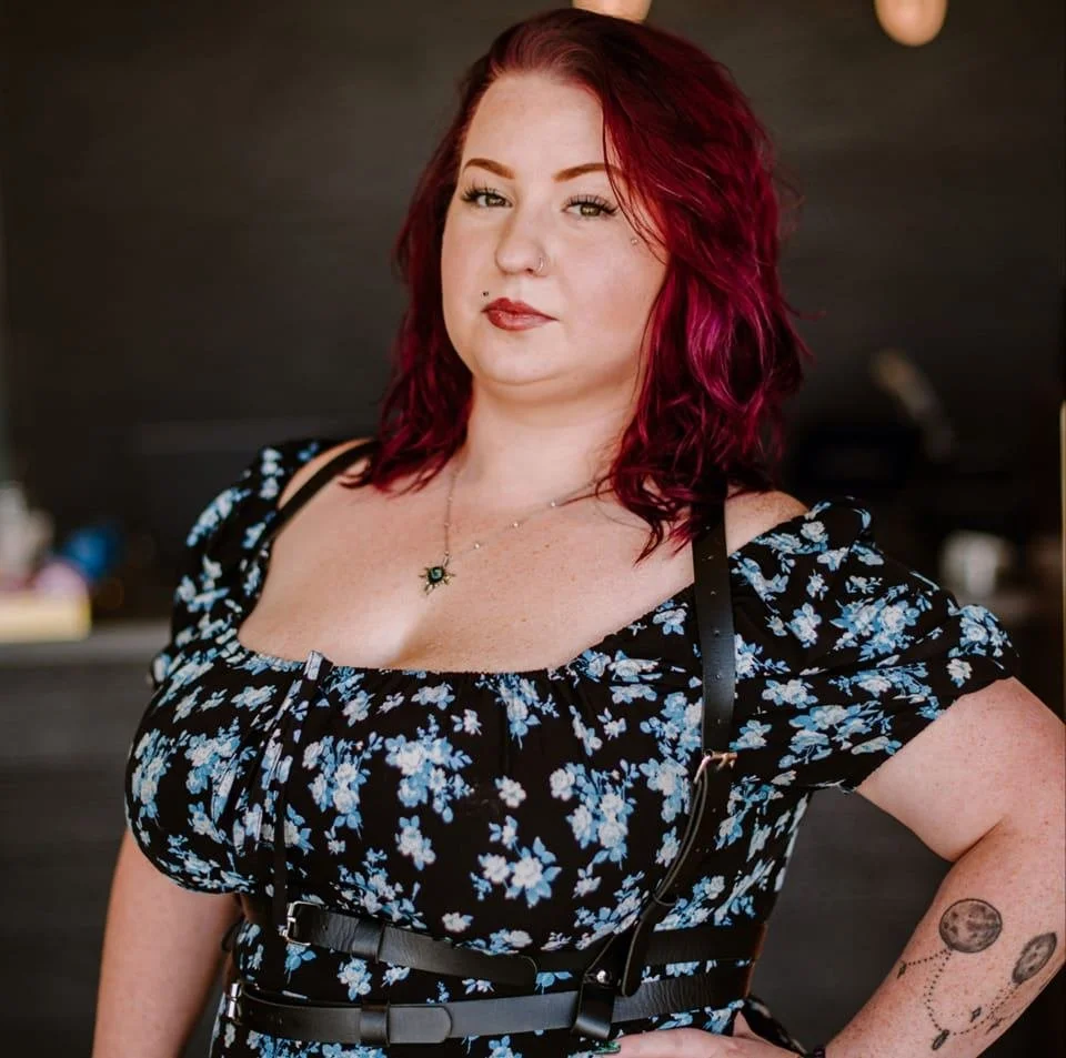 A woman with red hair and tattoos on her arm wearing a black floral dress and a necklace, standing indoors with a dark background.