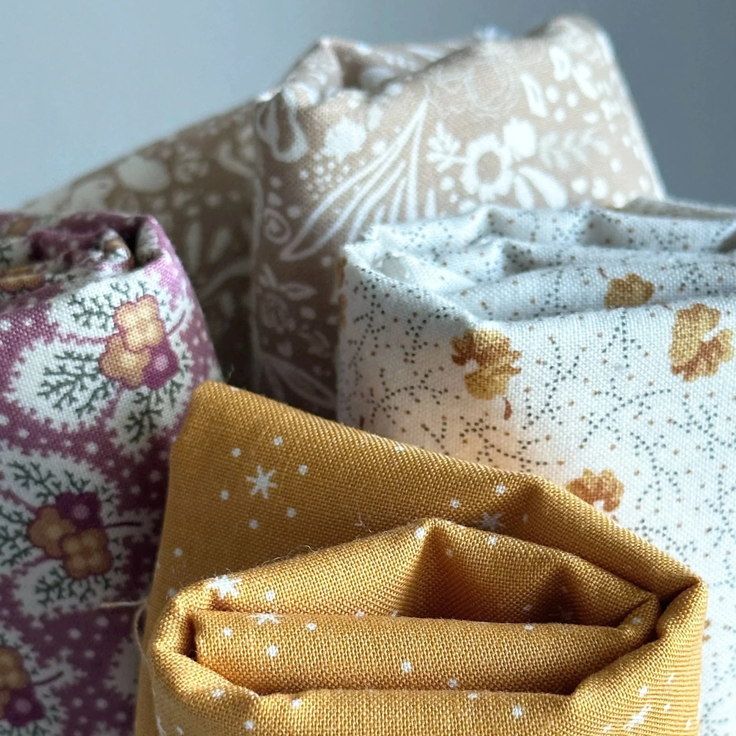 Folded cotton print fabrics in warm tones, including mustard, cream, beige, and pink floral patterns.