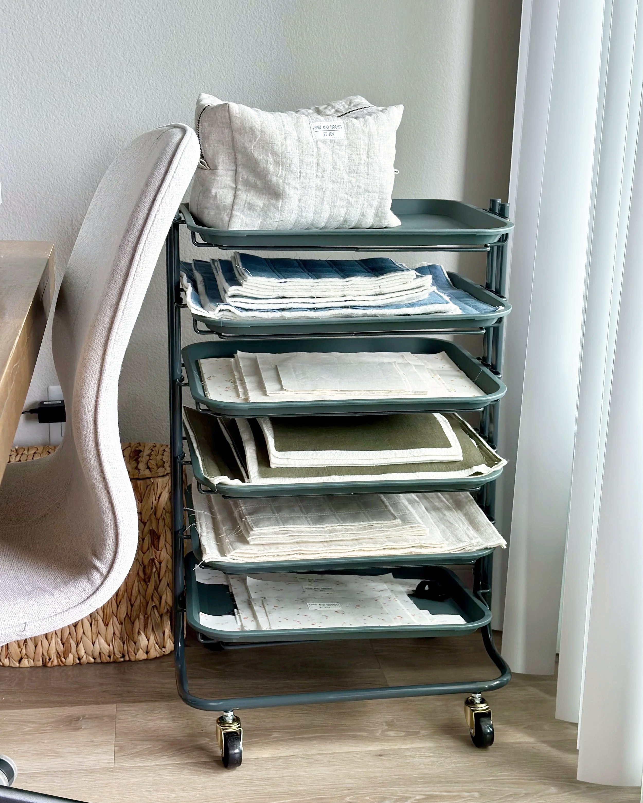 Rolling teal utility cart with wire trays holding stacks of folded quilted linen fabric in oat, navy stripe, olive, and cream beside a sewing chair