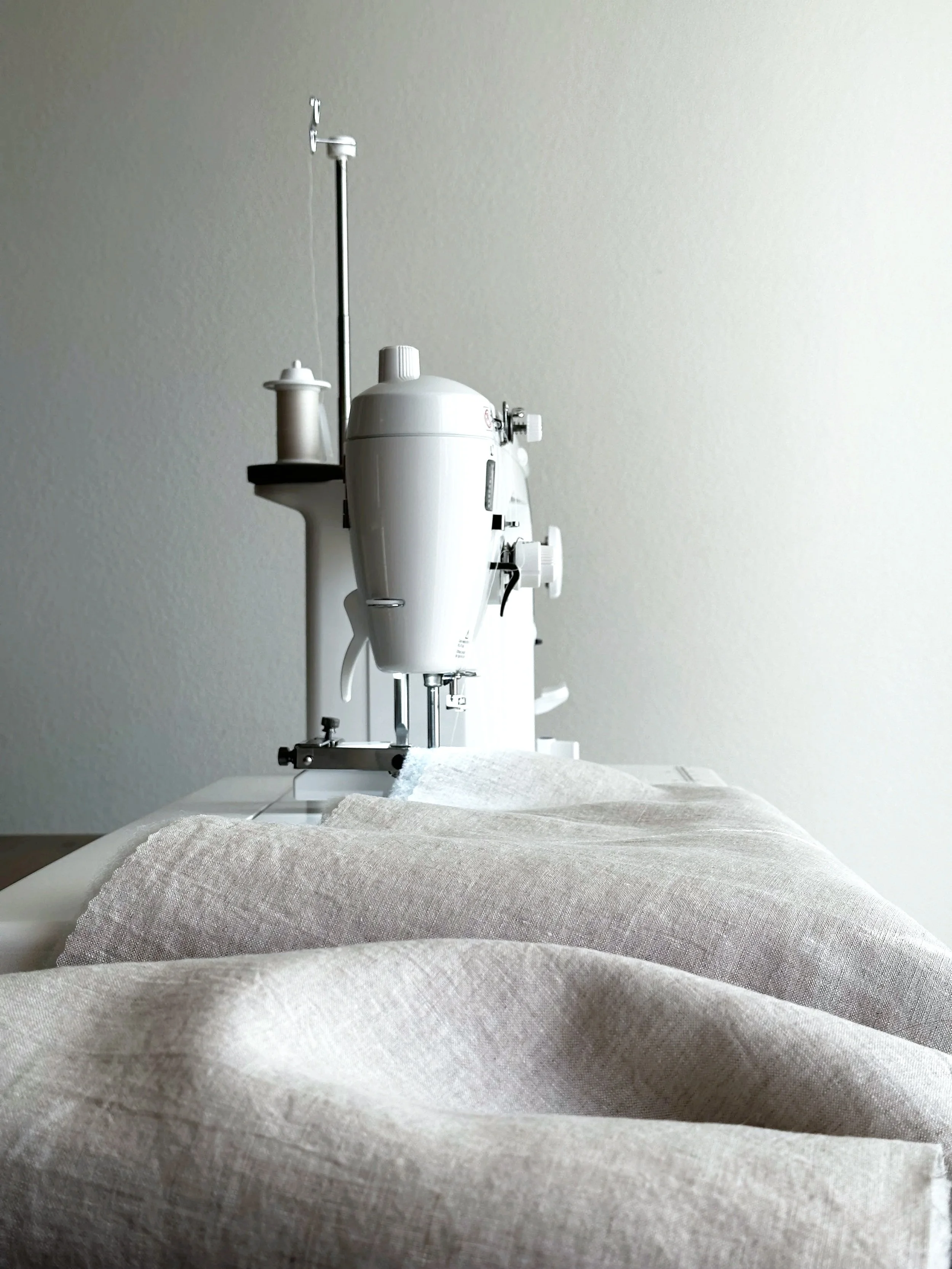 A sewing machine with beige fabric in front of it, positioned on a table against a plain light-colored wall.