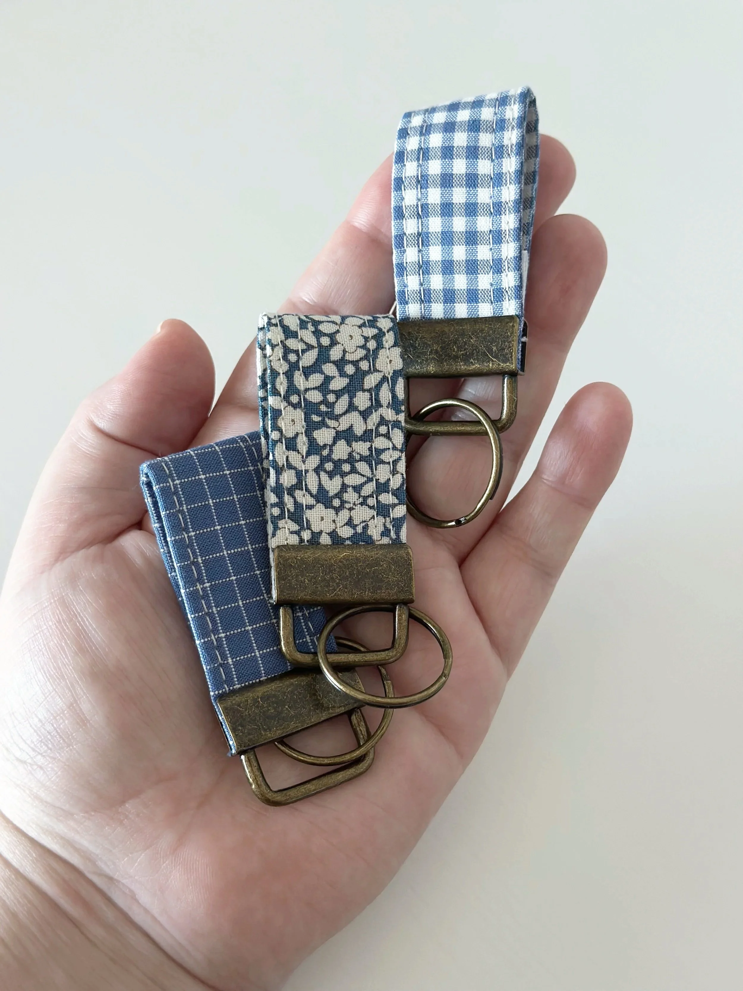 Hand holding three fabric key fobs in blue gingham, cream floral, and blue plaid with brass hardware