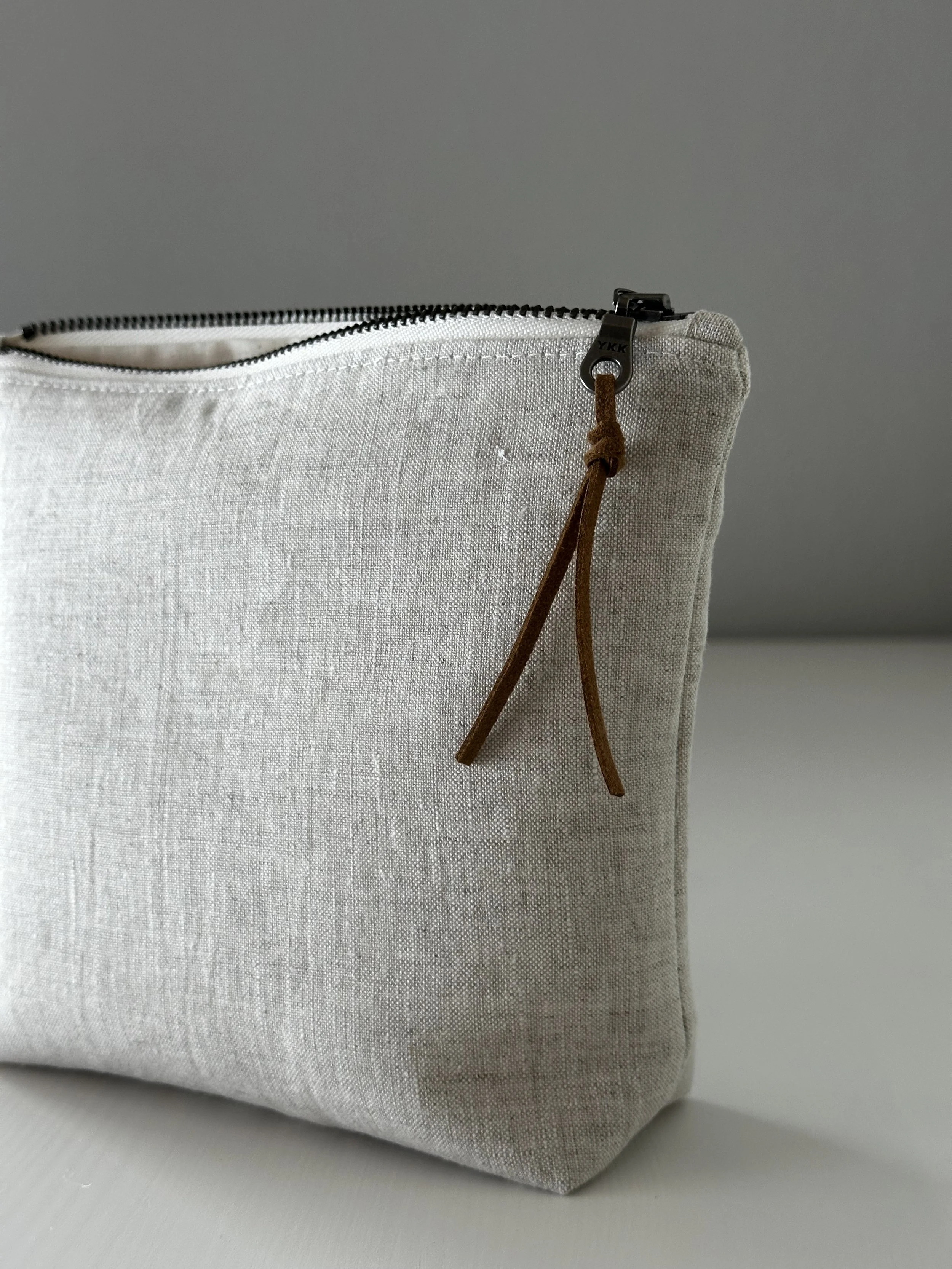 Light gray fabric zippered pouch with a brown leather pull tab.