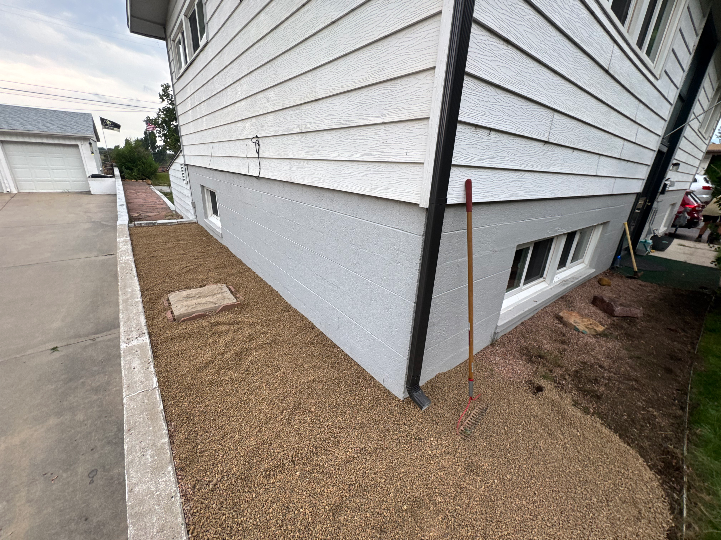 Side yard of a house with freshly laid gravel, a black downspout, and a rake leaning against the corner.