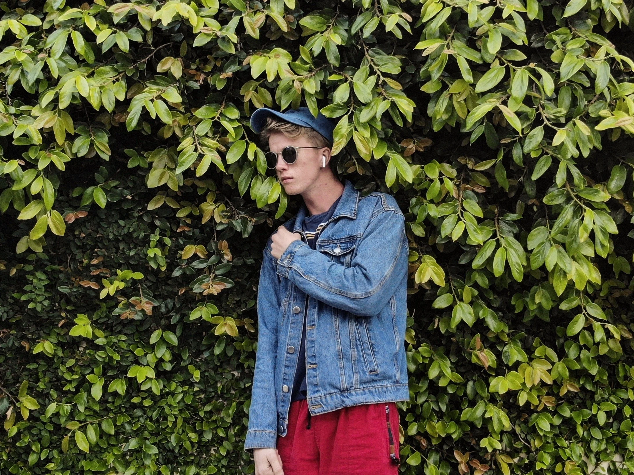 A young person standing in front of a dense green leafy hedge, wearing sunglasses, a blue baseball cap, a blue denim jacket, and red shorts.