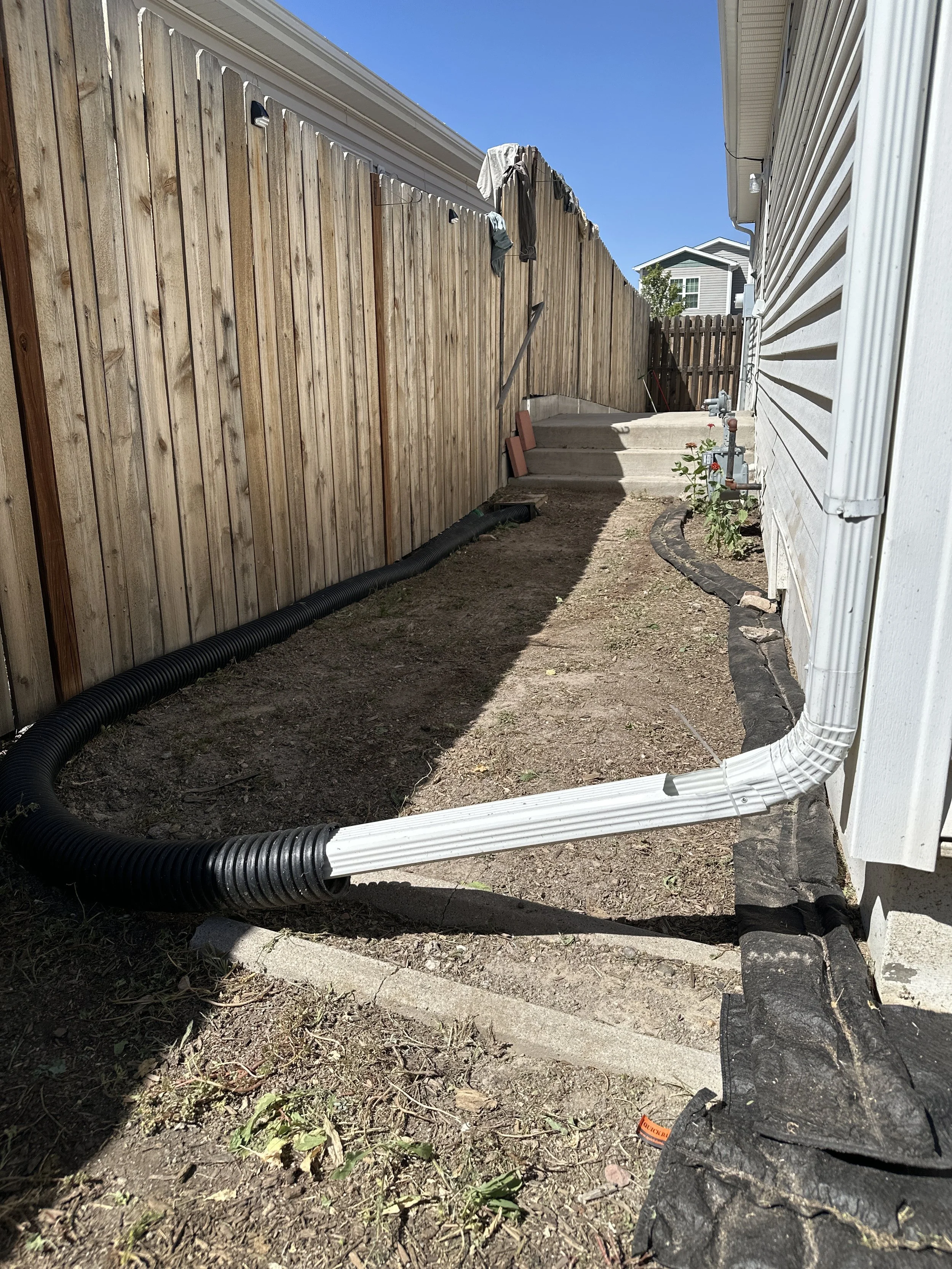 Partially landscaped backyard with a wooden fence on the left, a white house siding on the right, and dirt ground. There are black and white drainage pipes along the ground, a small patch of plants, and steps in the background leading to a higher lev