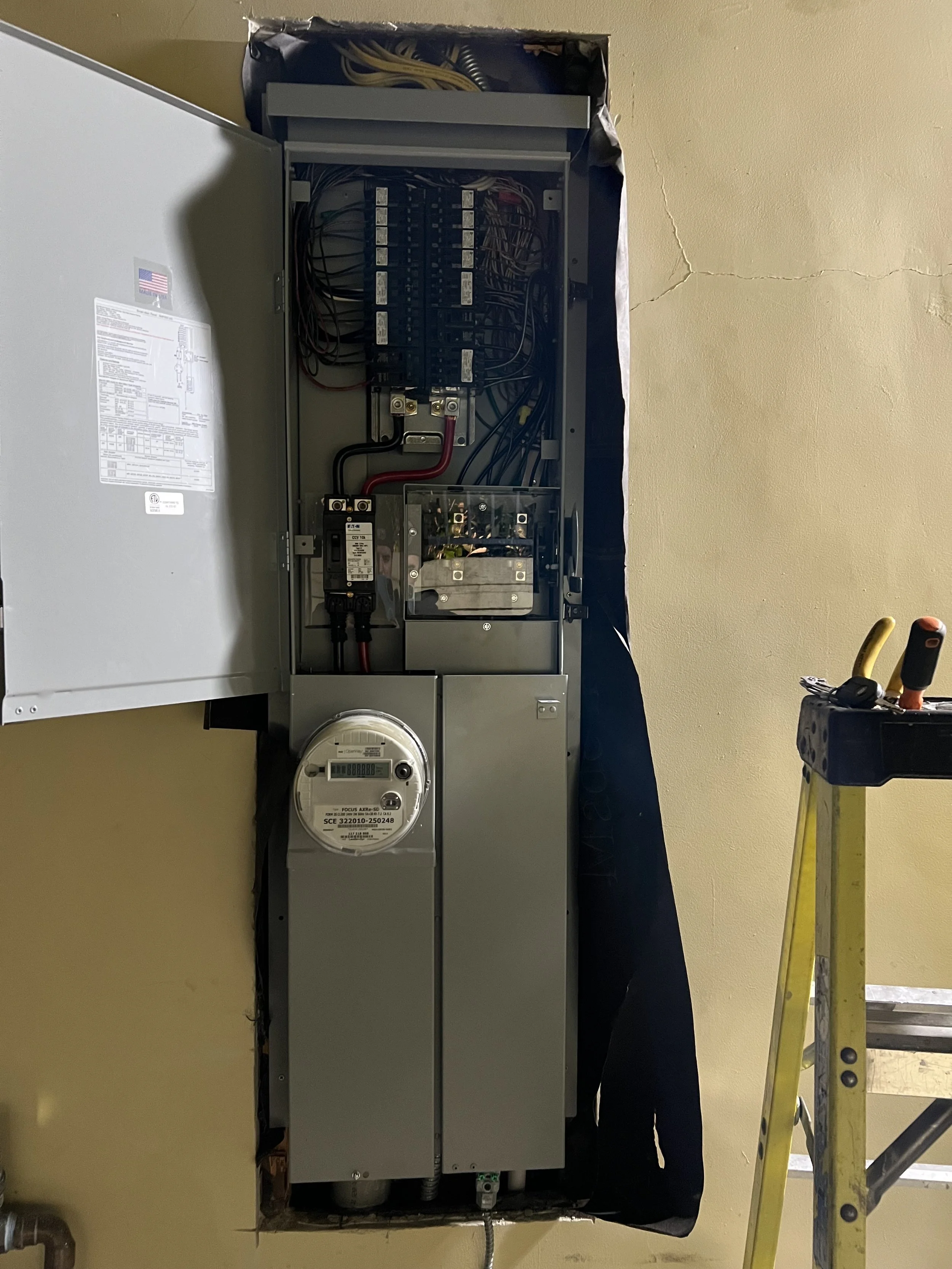 Electrical panel with open door revealing wiring, circuit breakers, a meter, and a connection to a utility outlet, with a yellow ladder and some tools beside it.