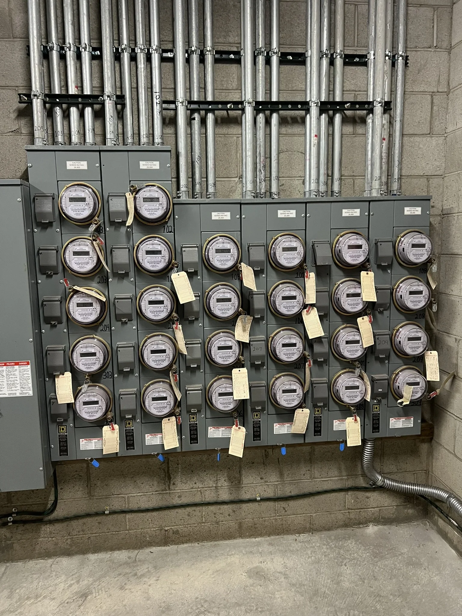 Multiple electric meters mounted on a gray electrical panel with various tubes and wires above and below.
