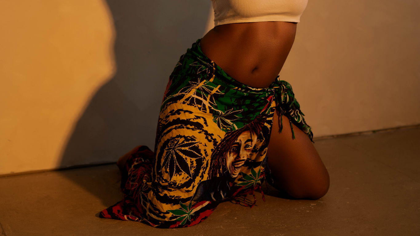 A person kneeling on the floor, wearing a skirt with vibrant Rastafarian colors and patterns, including a portrait of Bob Marley and marijuana leaf designs, with a white crop top and a tan background.
