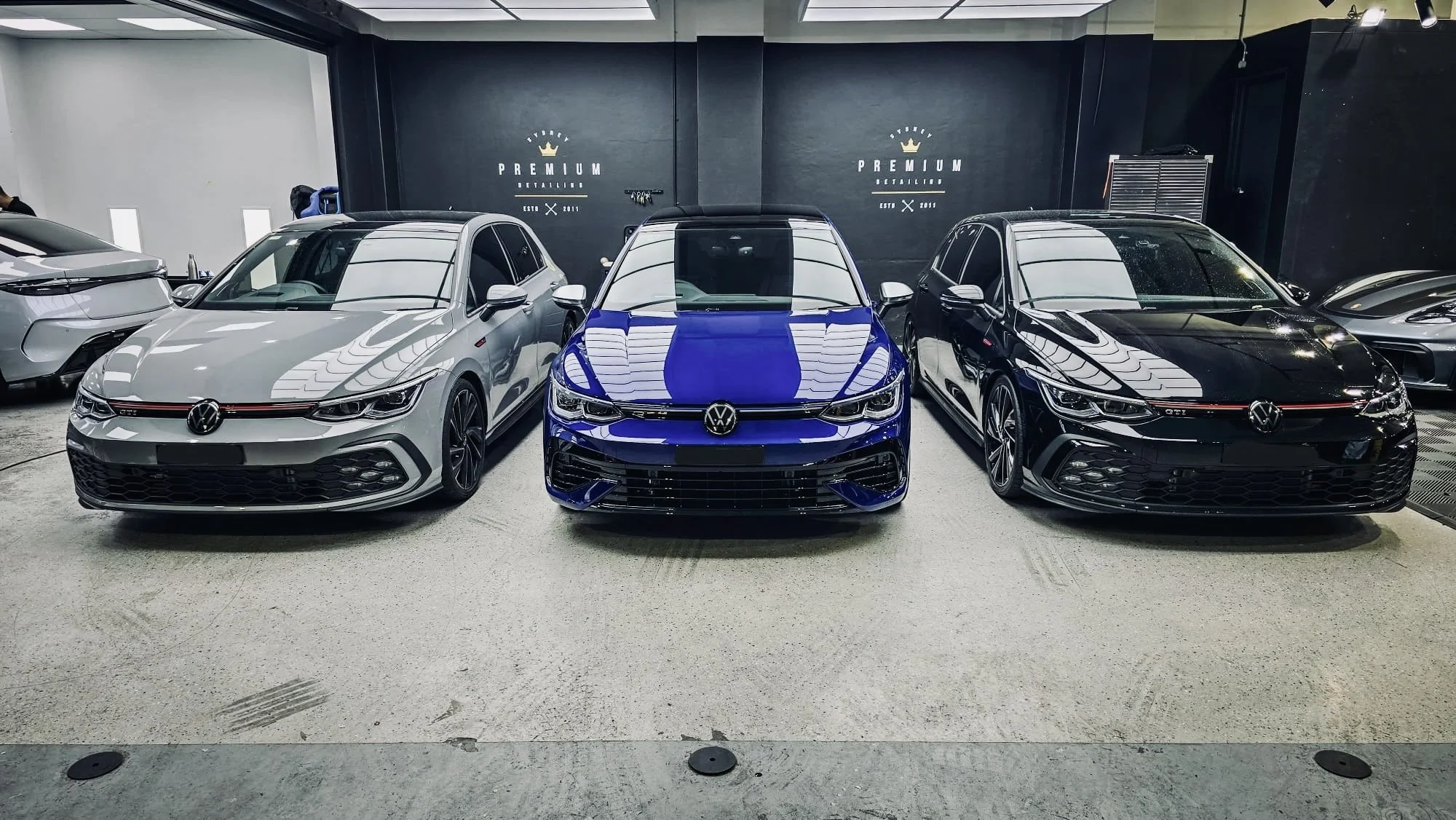 Three Volkswagen Golf GTI cars, one gray, one blue, and one black, parked side by side in a showroom.